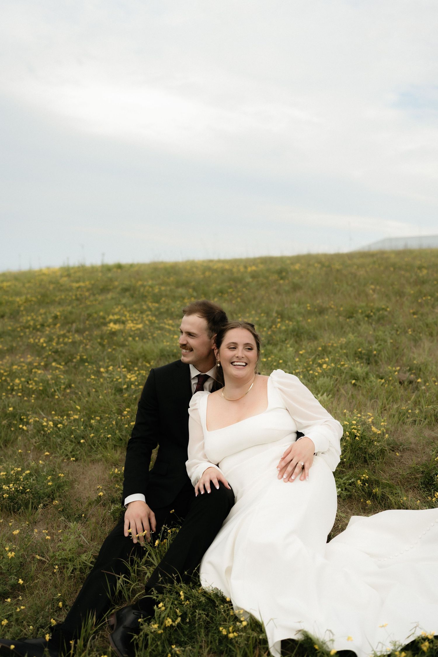 A joyful bride in a white gown and groom in black suit sit together on a flower-filled hillside under a cloudy sky.