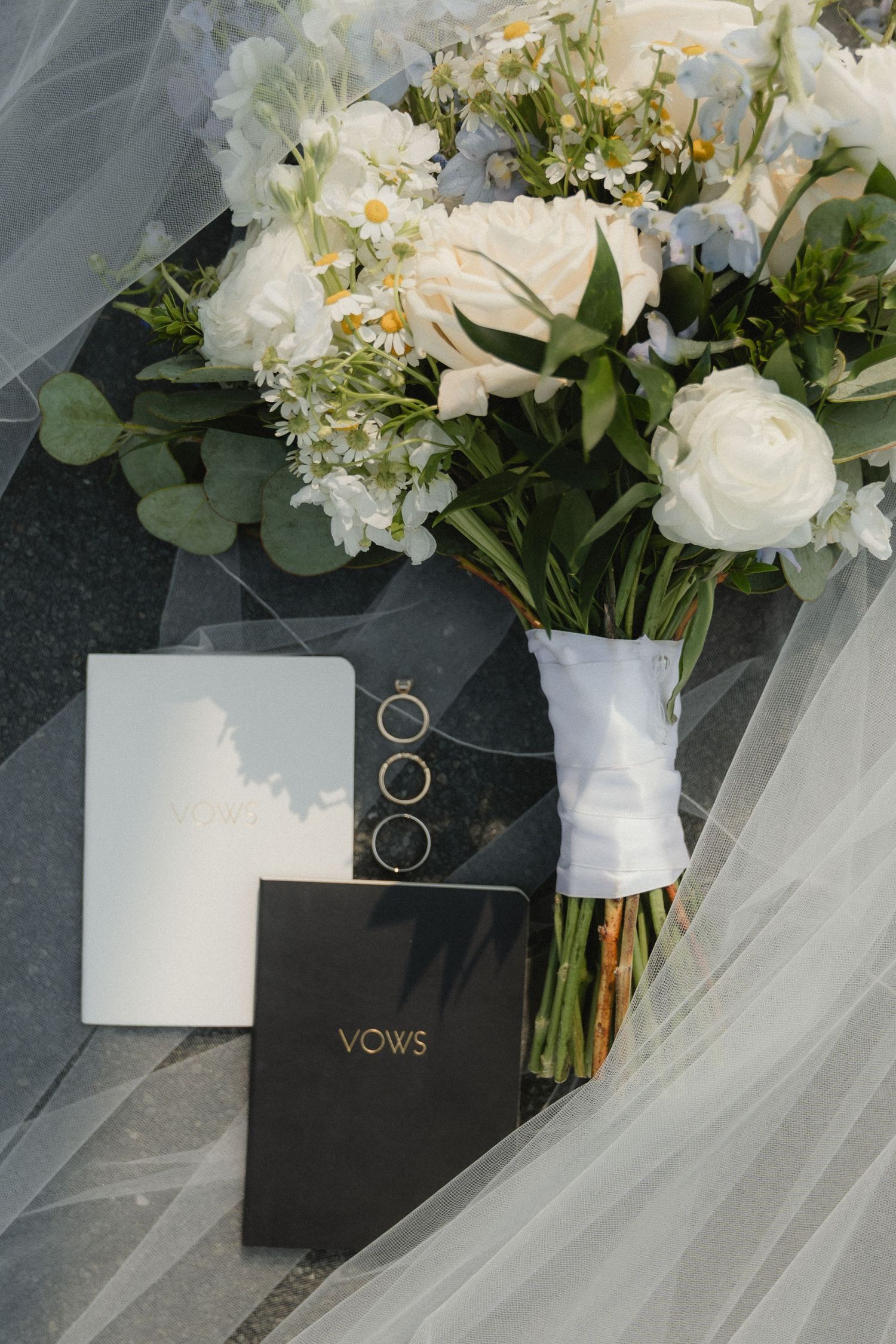 Wedding flat lay with white floral bouquet, vow books, and gold rings draped with elegant bridal veil.