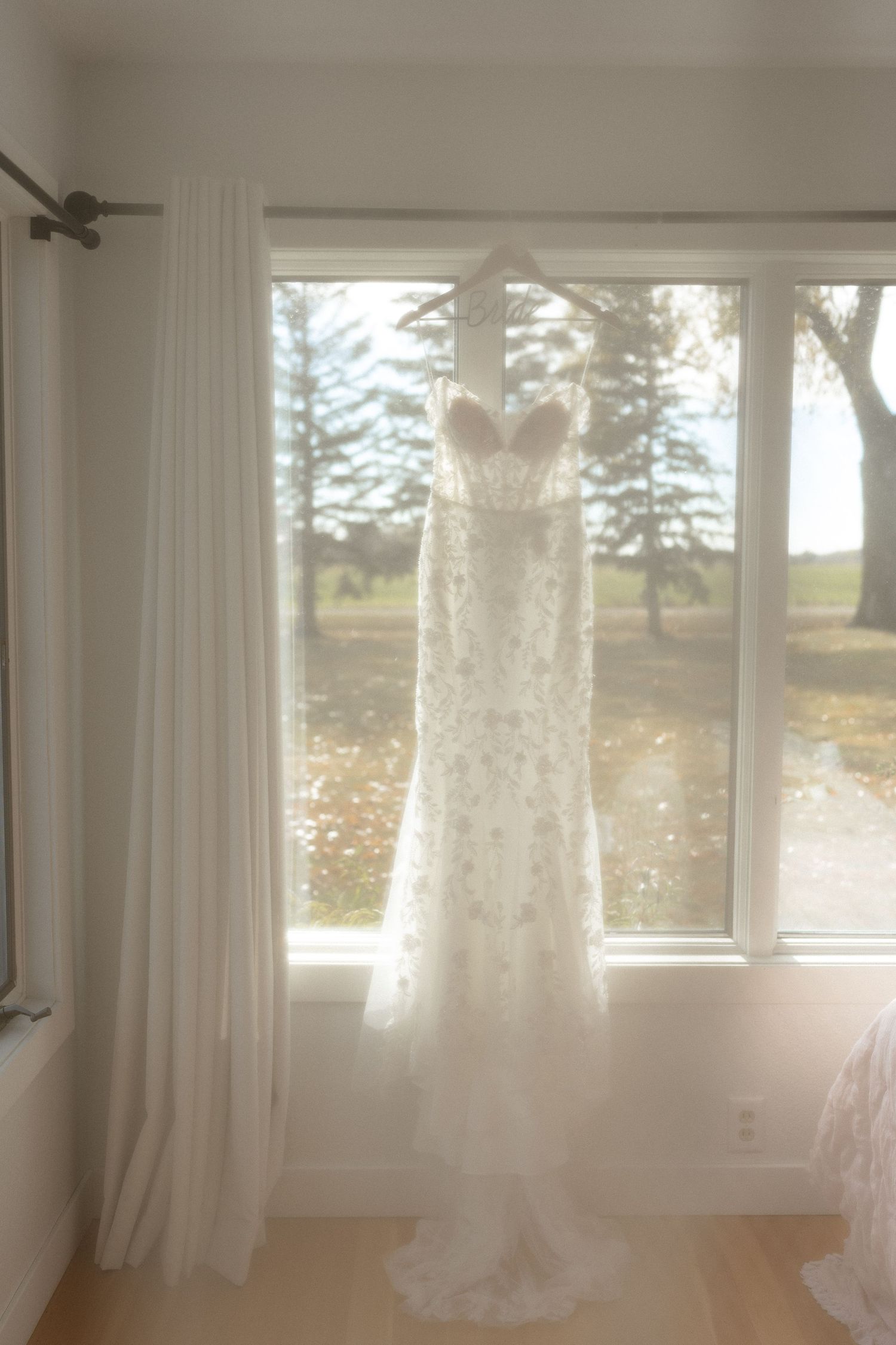 A lace wedding dress hangs in front of sunlit windows creating an ethereal silhouette.