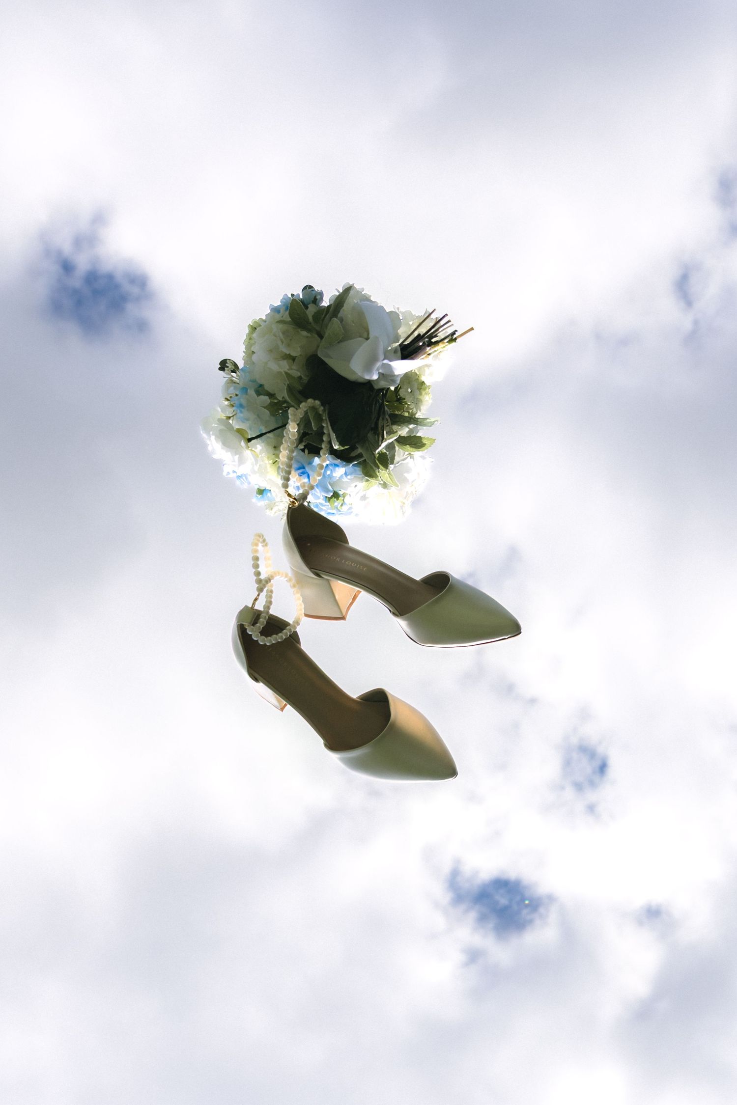 Bridal heels and floral bouquet with blue flowers floating against a cloudy sky backdrop.