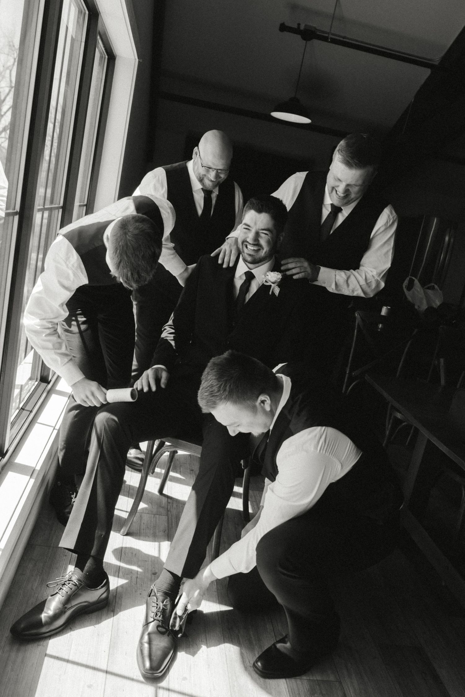Black and white photo of a groom laughing as groomsmen help him get dressed before the wedding ceremony.