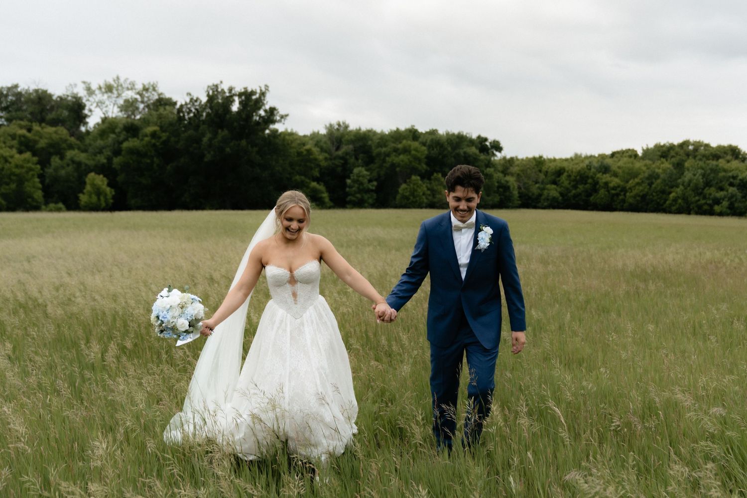 Bride and groom walk hand-in-hand through a lush green field, bride holding a blue bouquet in a white gown.