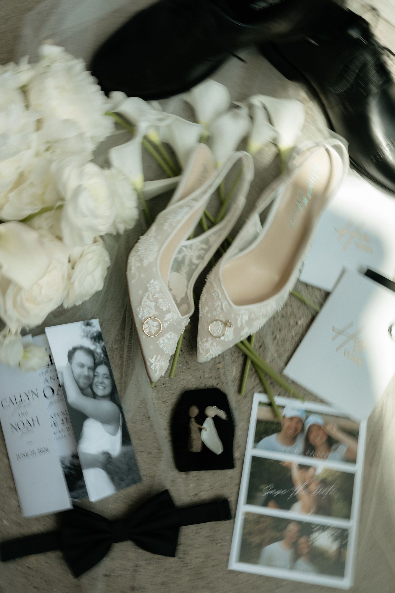 Elegant bridal flat lay with lace heels, white rose bouquet, wedding invitations, bow tie, and couple photos on neutral background.