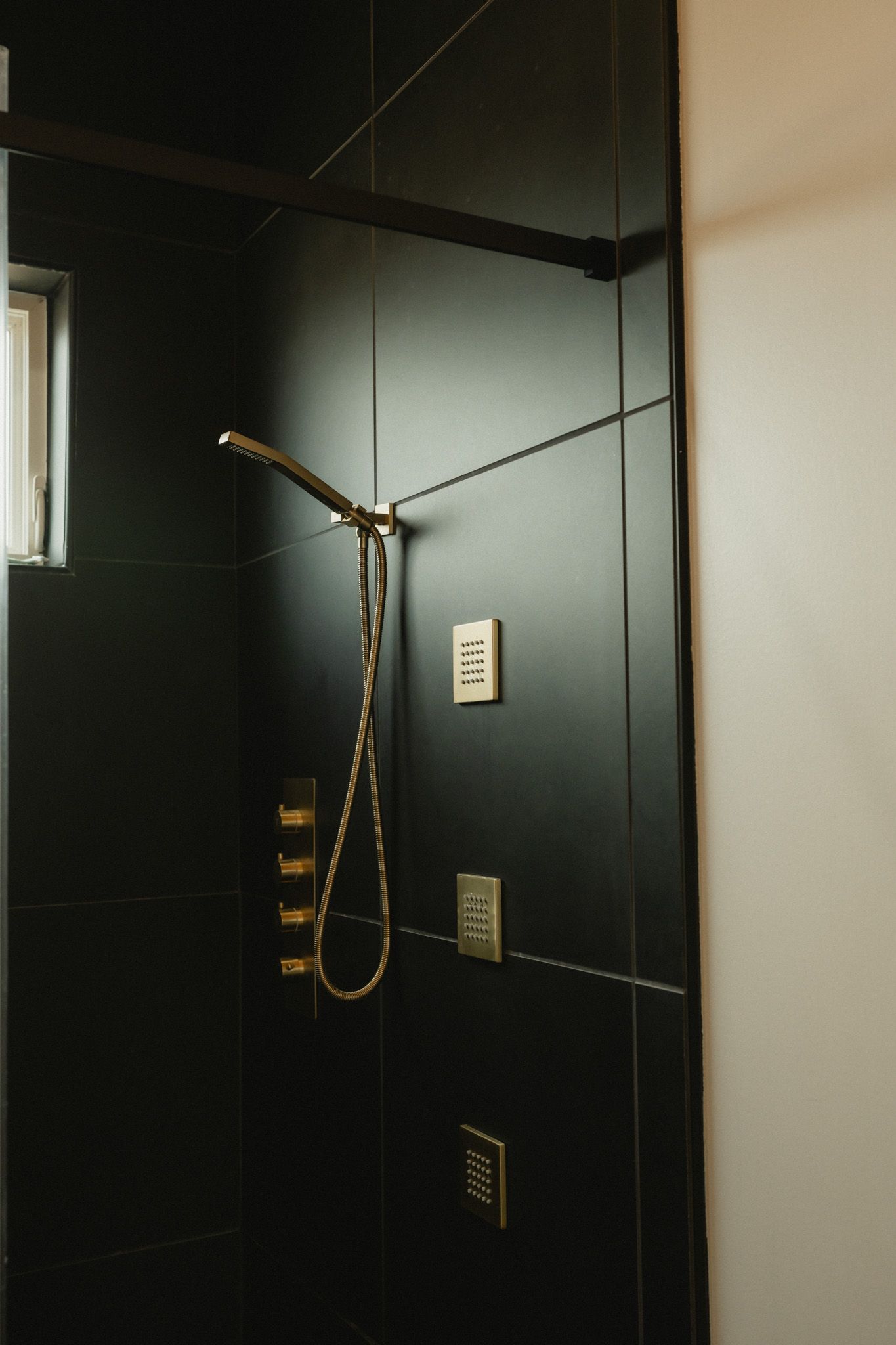 Modern shower with black tiles and gold fixtures, featuring a handheld showerhead and sleek wall-mounted controls.