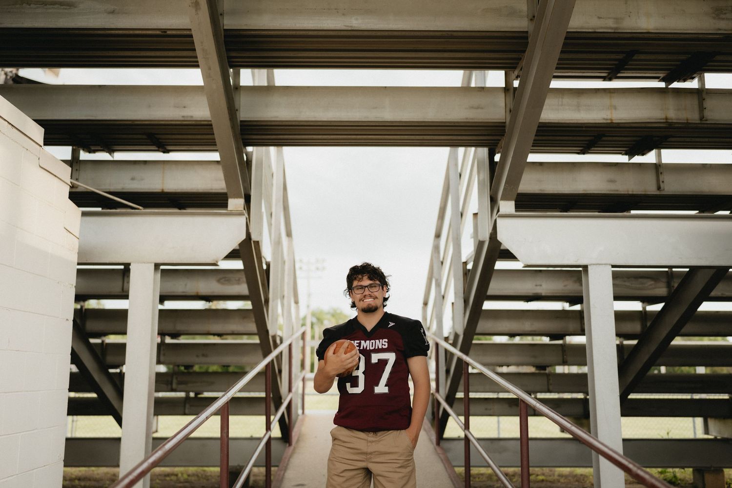 Young football player wearing jersey number 37 stands confidently on stadium bleachers holding a ball.