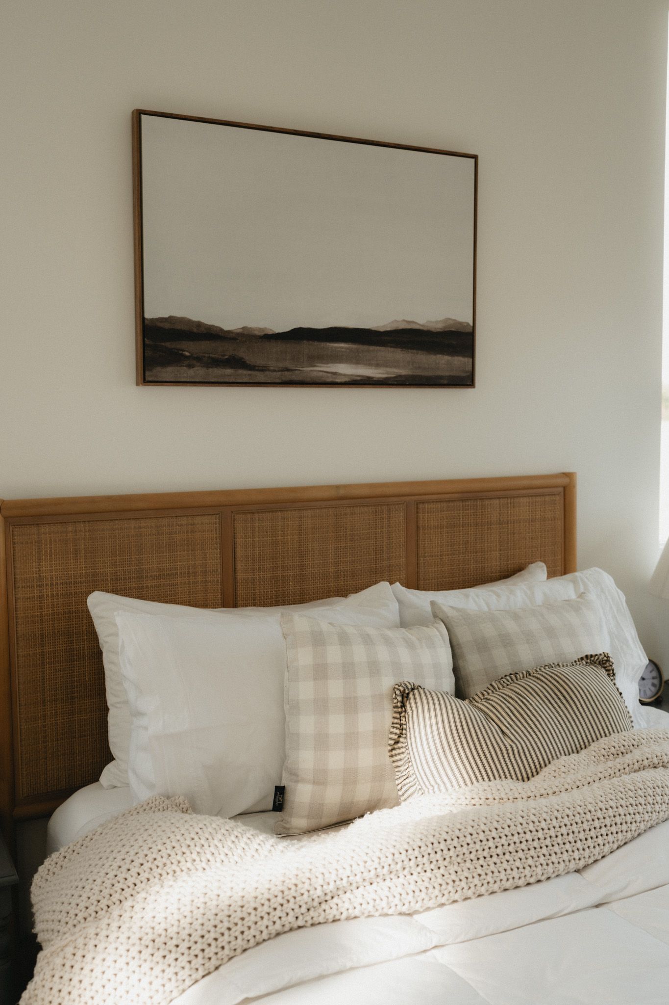 Neutral bedroom with rattan headboard, layered pillows, knit blanket, and abstract landscape art above the bed.