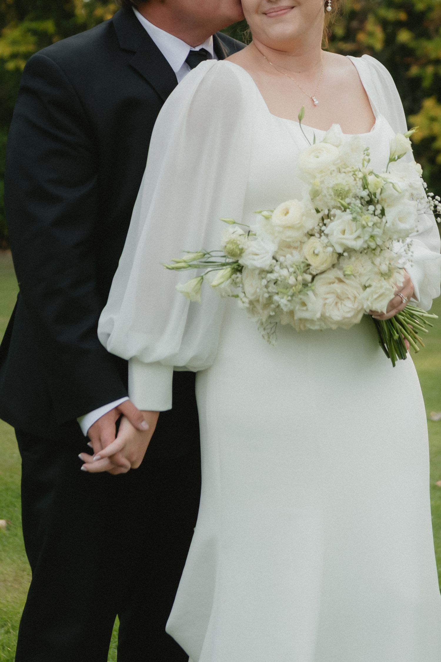 Bride in white gown with sheer sleeves holding bouquet of white roses, groom in black suit kissing her cheek outdoors.