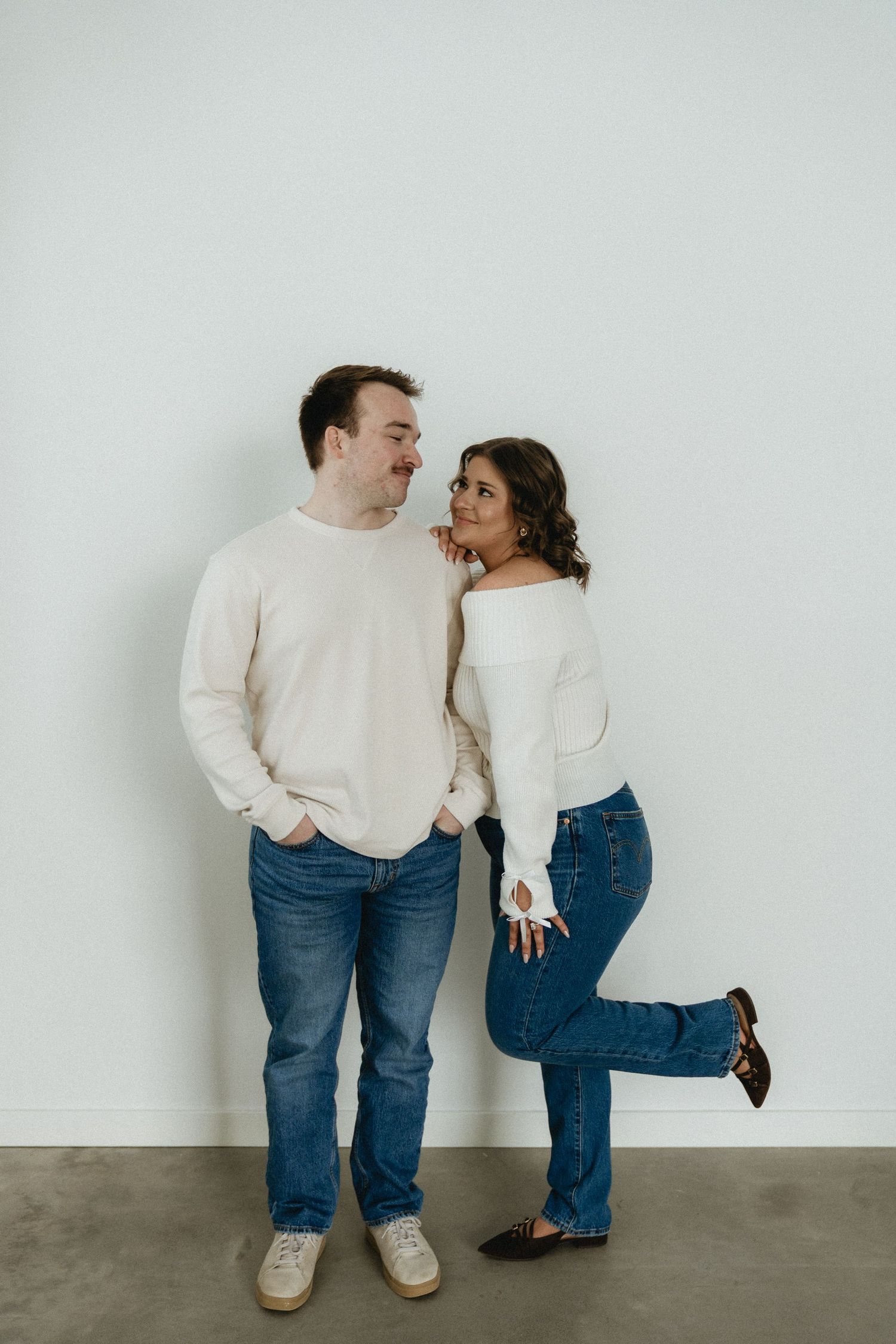 A couple in matching cream sweaters and jeans poses playfully against a minimalist white wall.