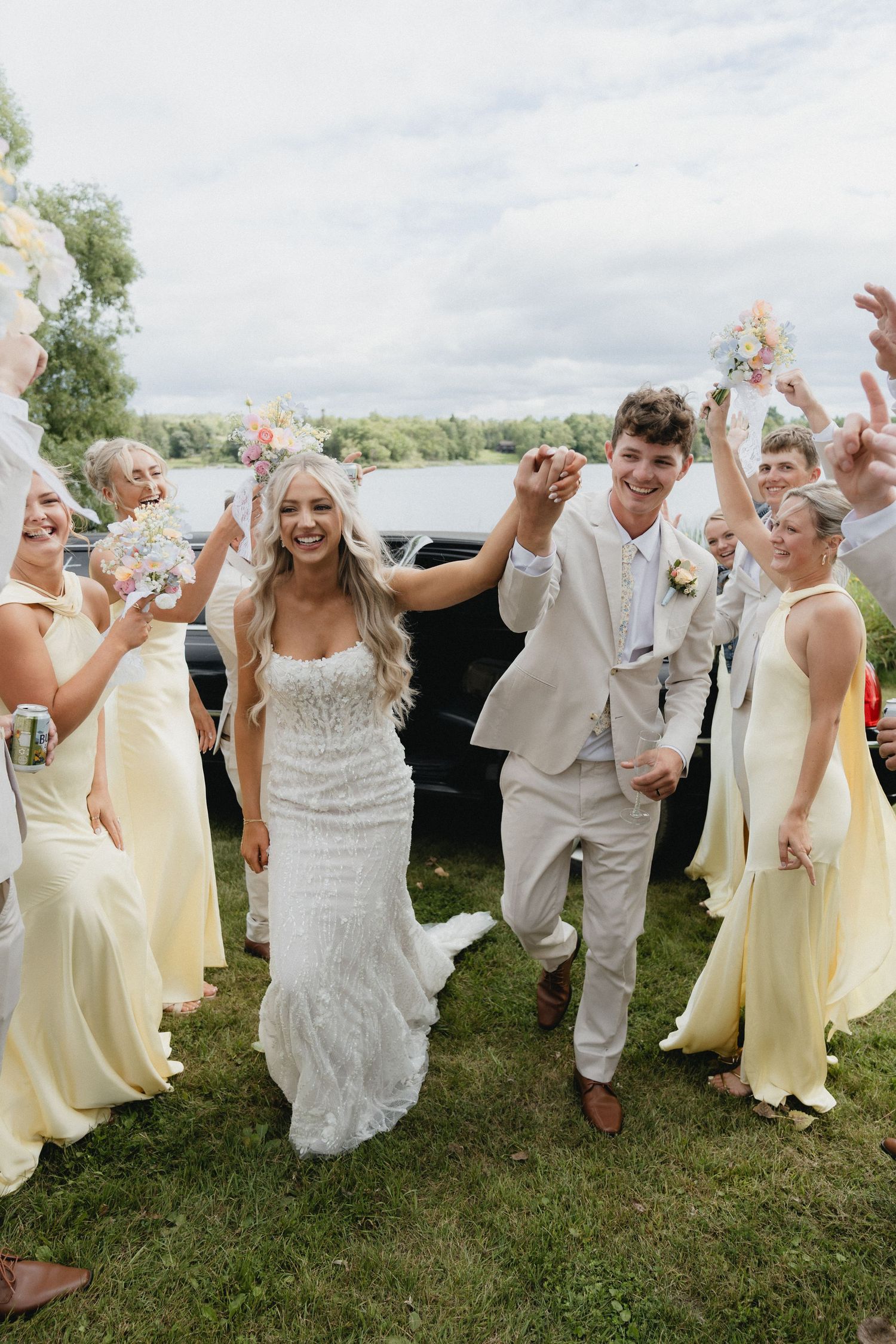 Wedding party in white and yellow attire celebrates joyfully on a grassy lawn under cloudy skies.