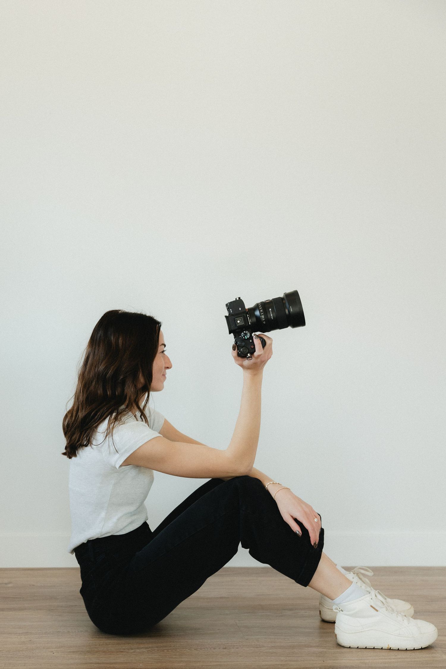 Photographer sitting on wooden floor in casual attire holding professional camera against white wall.