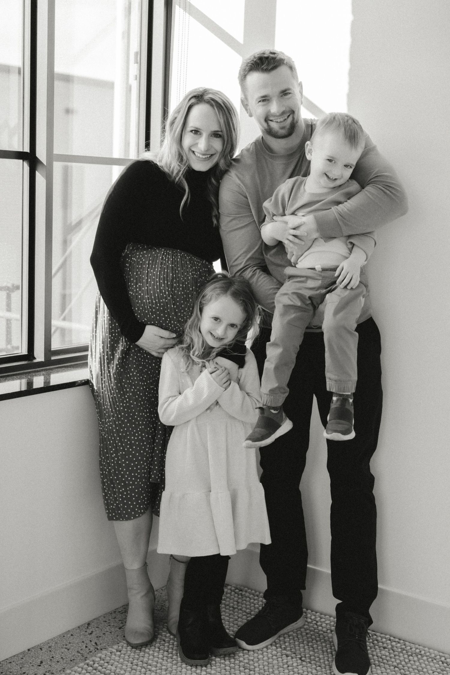 Black and white family portrait with pregnant mother, father holding toddler son, and young daughter smiling by window.