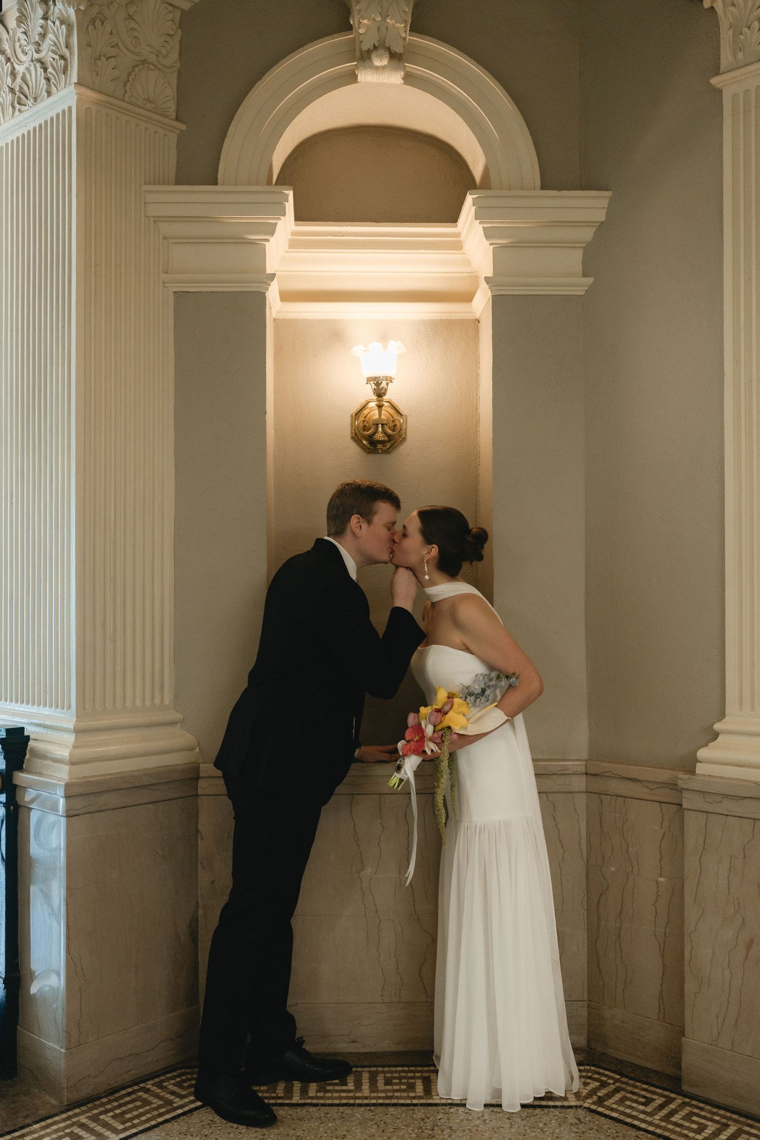 Bride and groom share a romantic kiss in an elegant arched alcove, bride holding colorful bouquet in white gown.