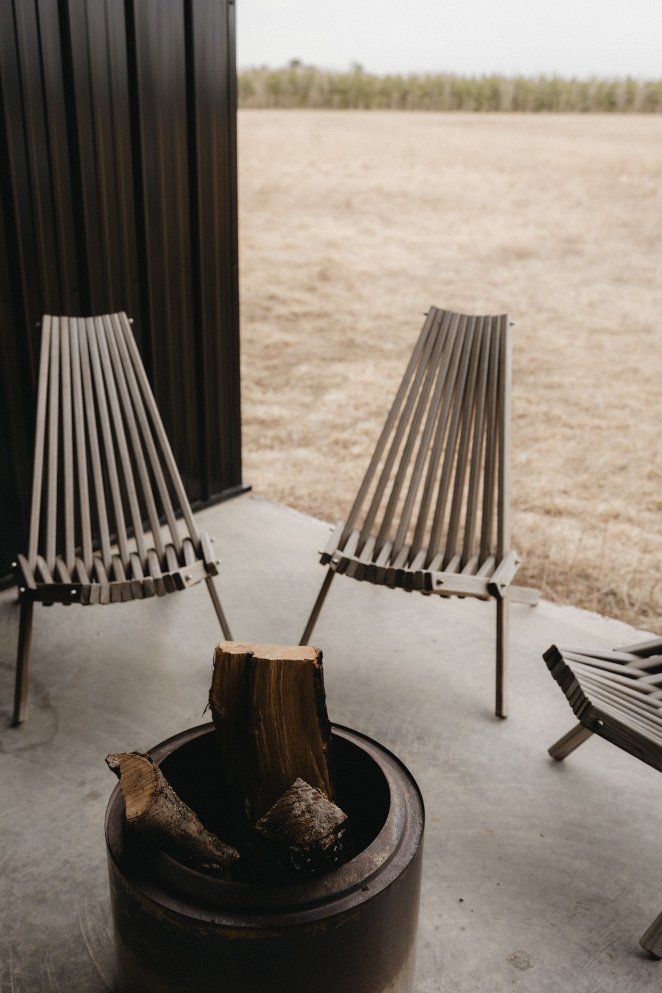 Modern slatted metal chairs on a concrete patio with a rustic fire pit holding logs, overlooking a dry grass field.