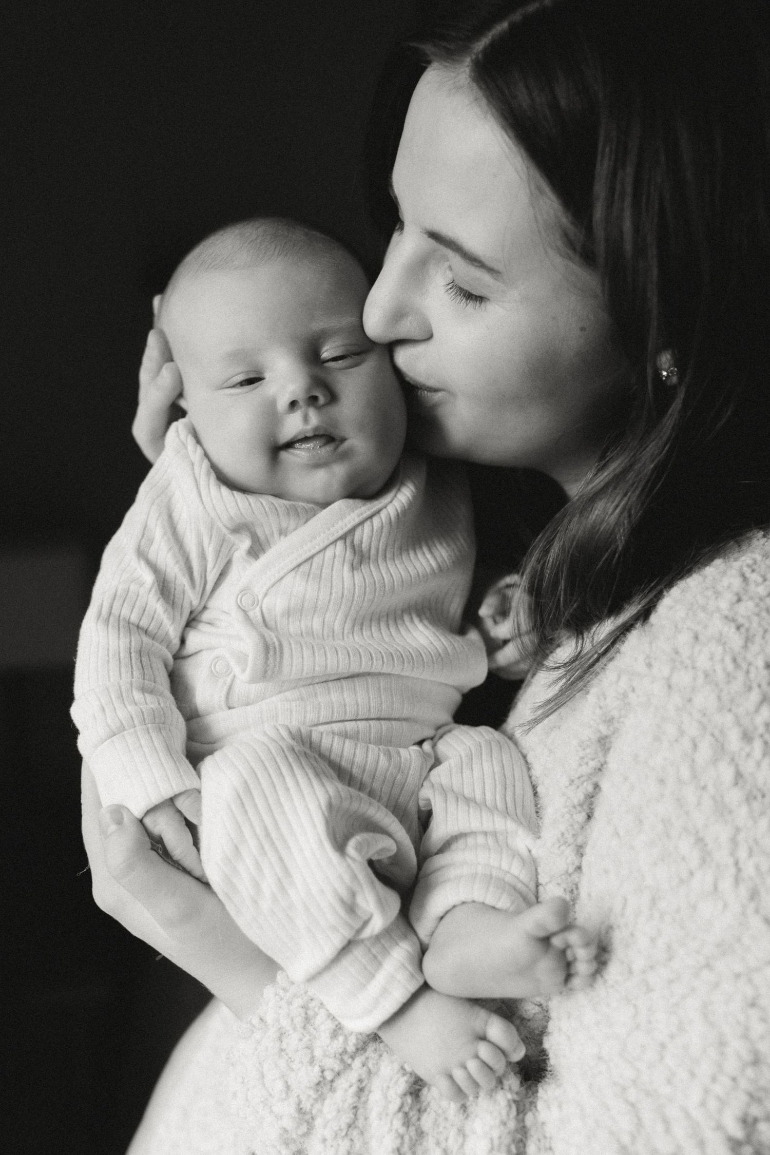 Black and white photo of a young mother tenderly kissing her newborn baby while cradling them in her arms.
