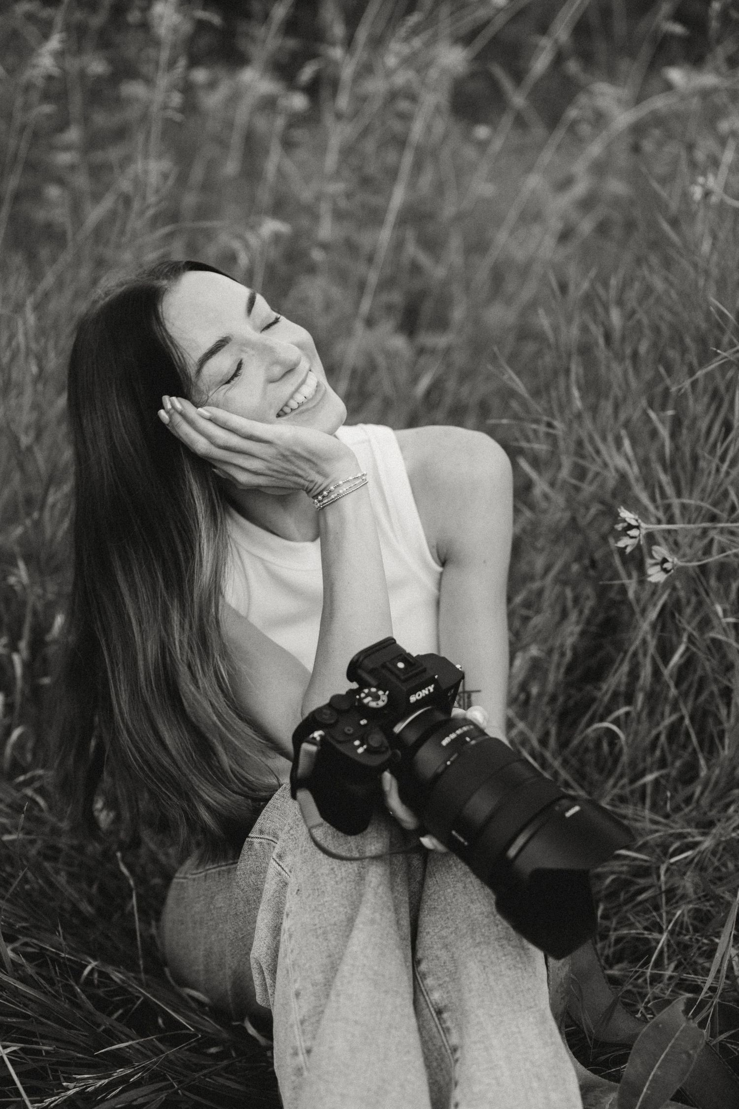 A photographer with long dark hair smiles while holding a professional camera in a field of tall grass.