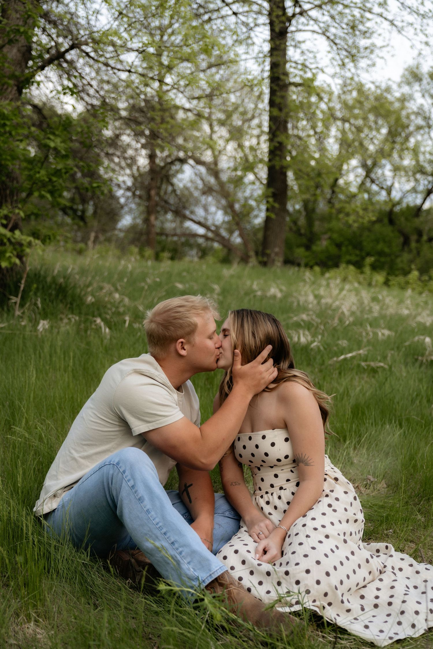 A couple shares a romantic kiss while sitting on green grass in a park, woman wearing a white polka dot dress.
