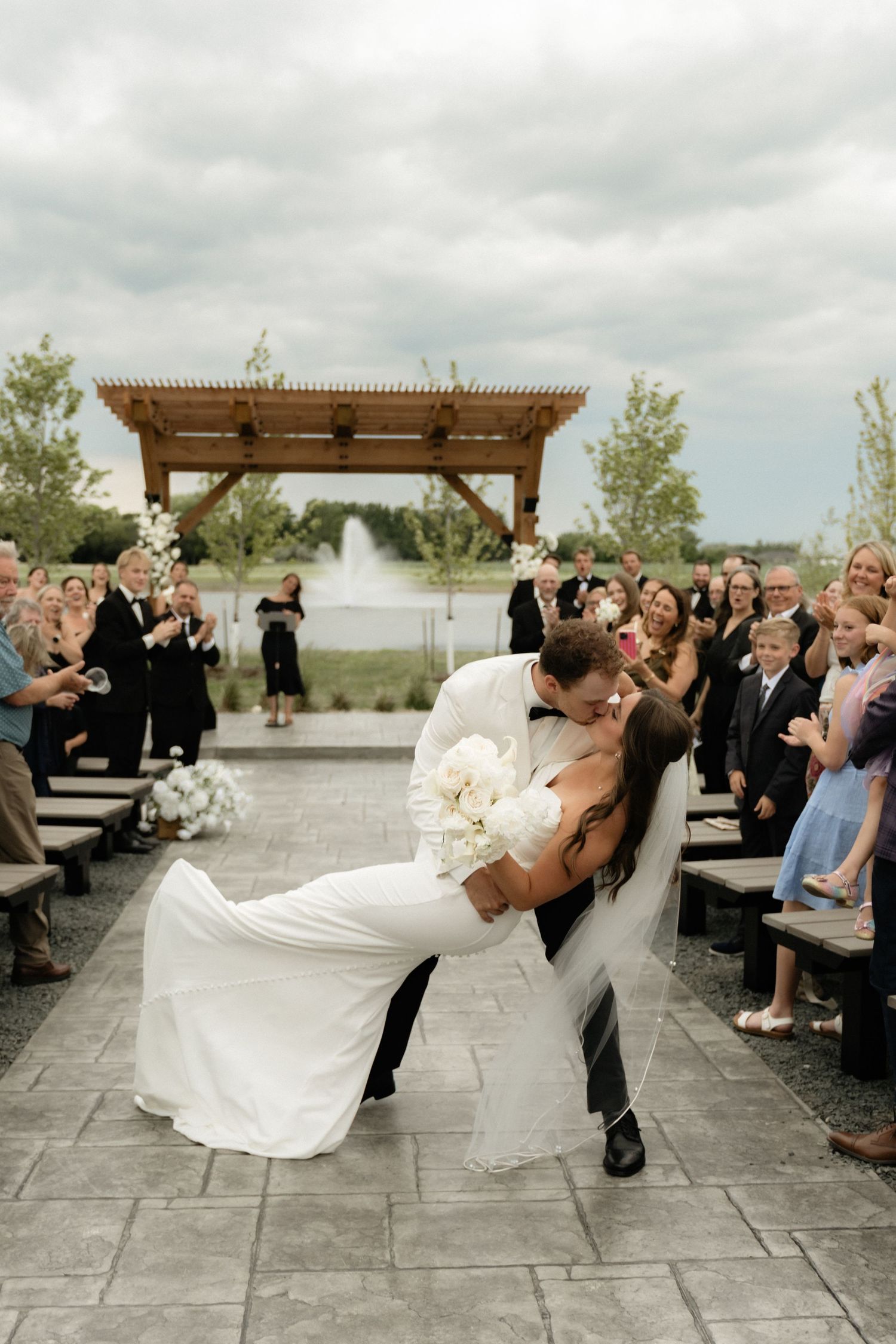Groom dips bride for a kiss at outdoor wedding ceremony with fountain backdrop and cheering guests lining the aisle.