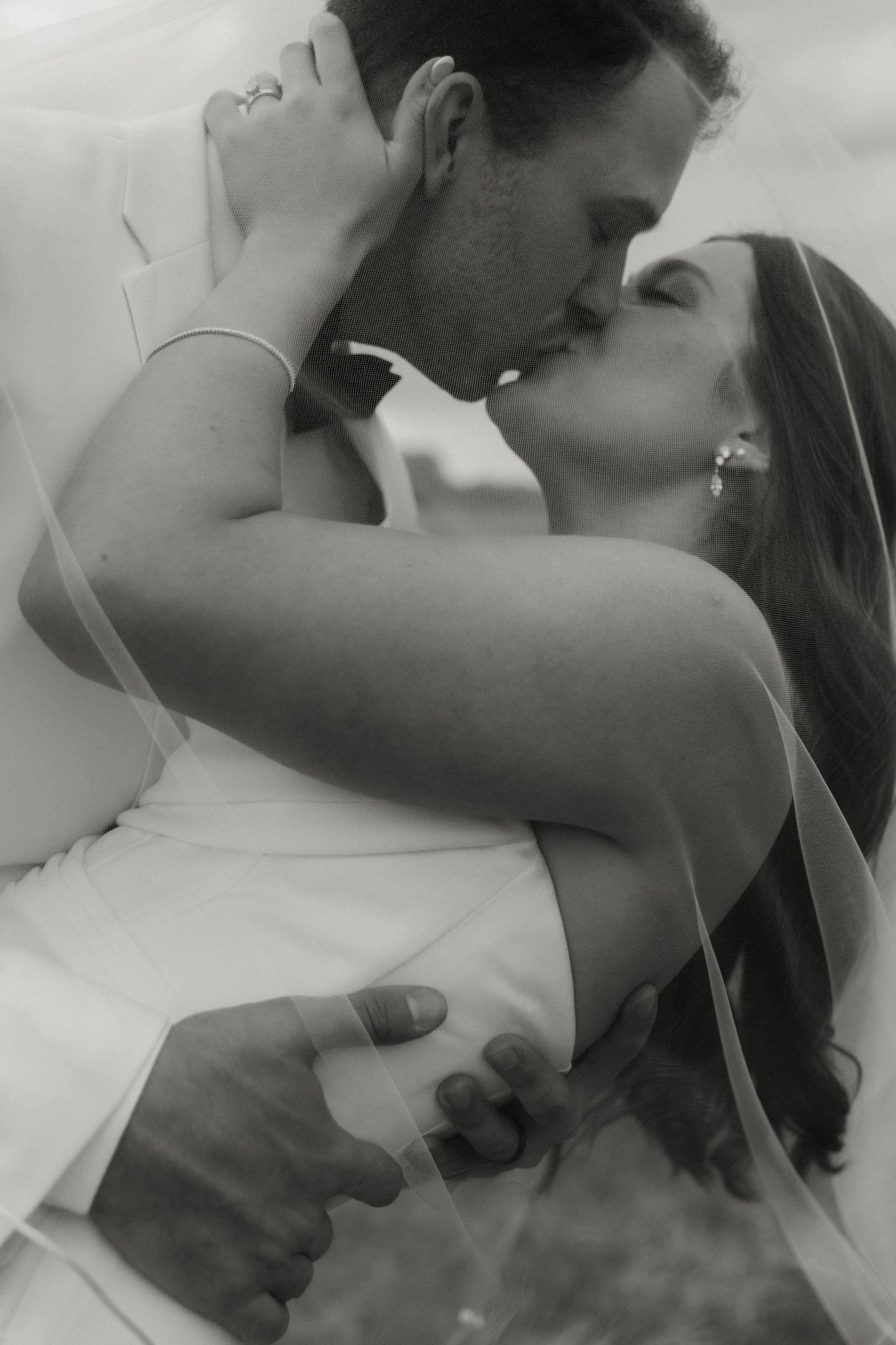 Black and white photo of a bride and groom sharing a romantic kiss under a flowing veil on their wedding day.