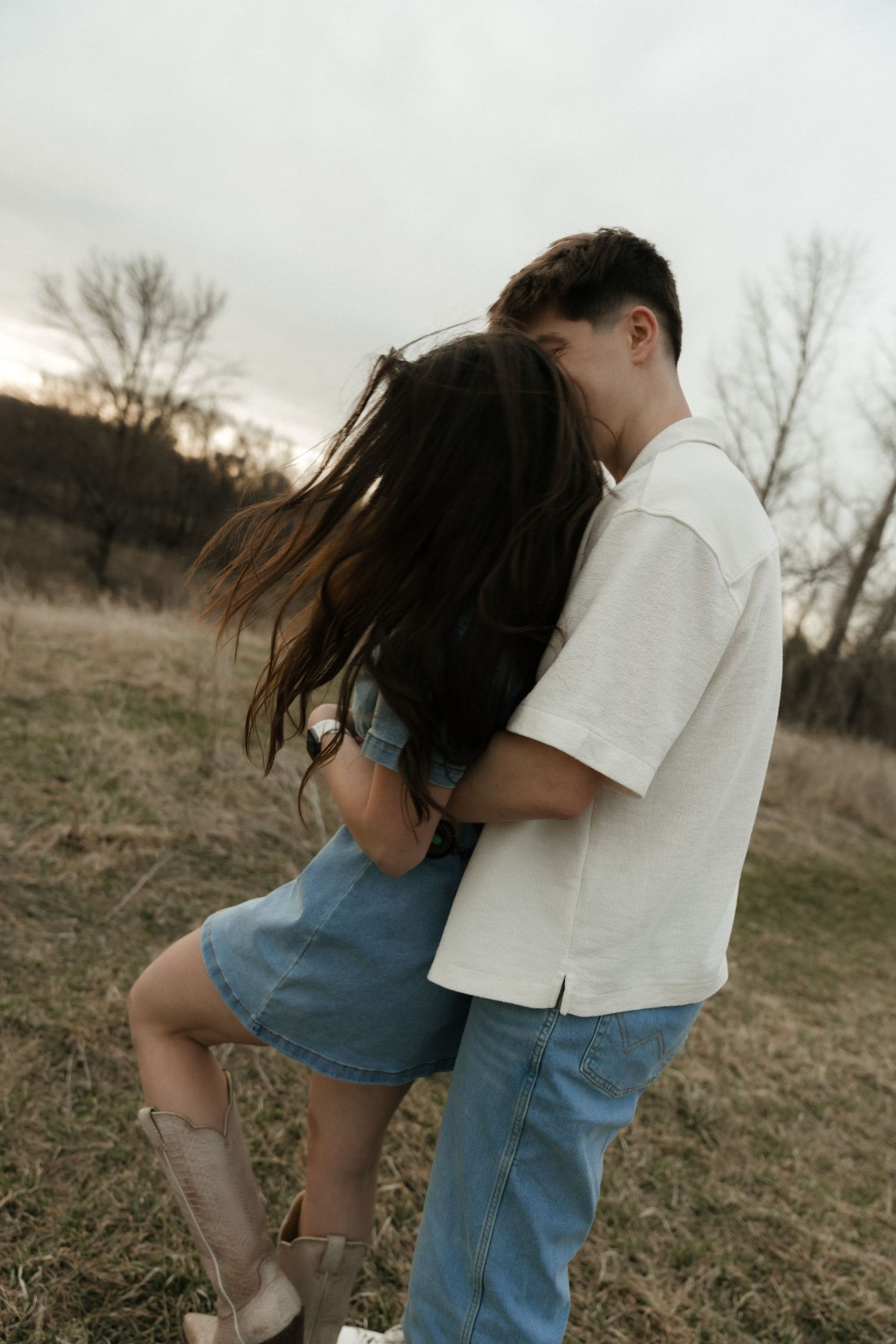 A couple embraces romantically in a field at dusk, the woman in a denim dress and cowboy boots, man in white shirt.