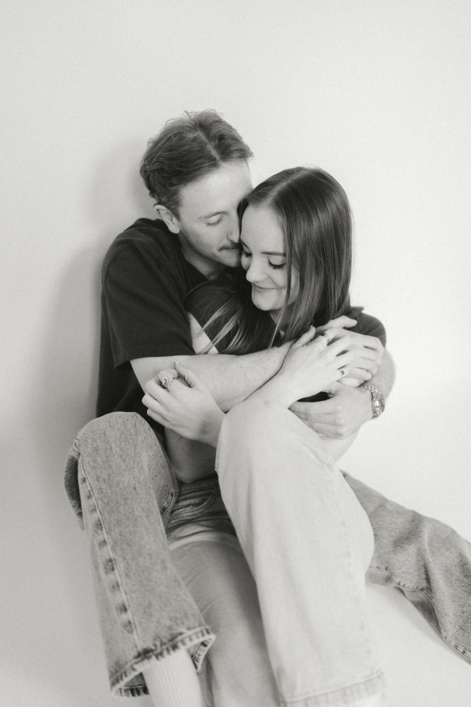 Black and white photo of a couple embracing tenderly, sitting against a white wall, sharing an intimate moment.