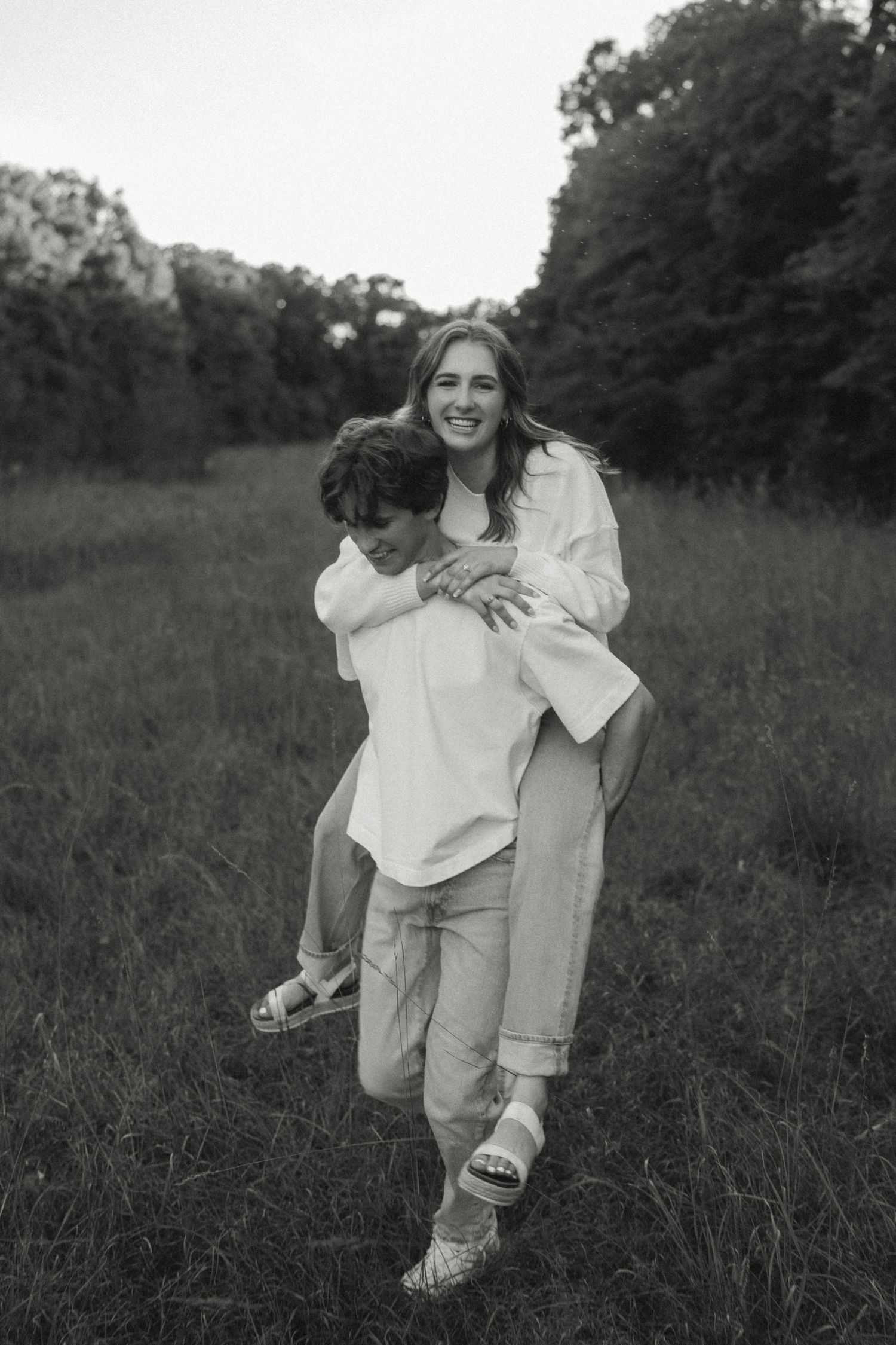 Black and white photo of a couple in a field, woman riding piggyback on man, both smiling, trees in background.