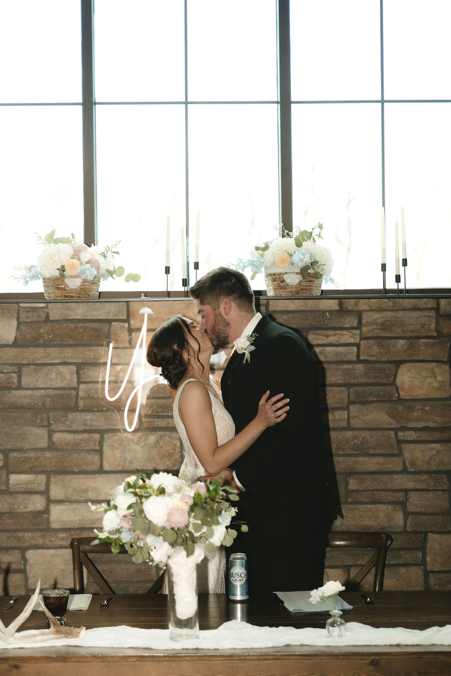 Bride and groom share first kiss at rustic stone wall sweetheart table with floral arrangements and candles.