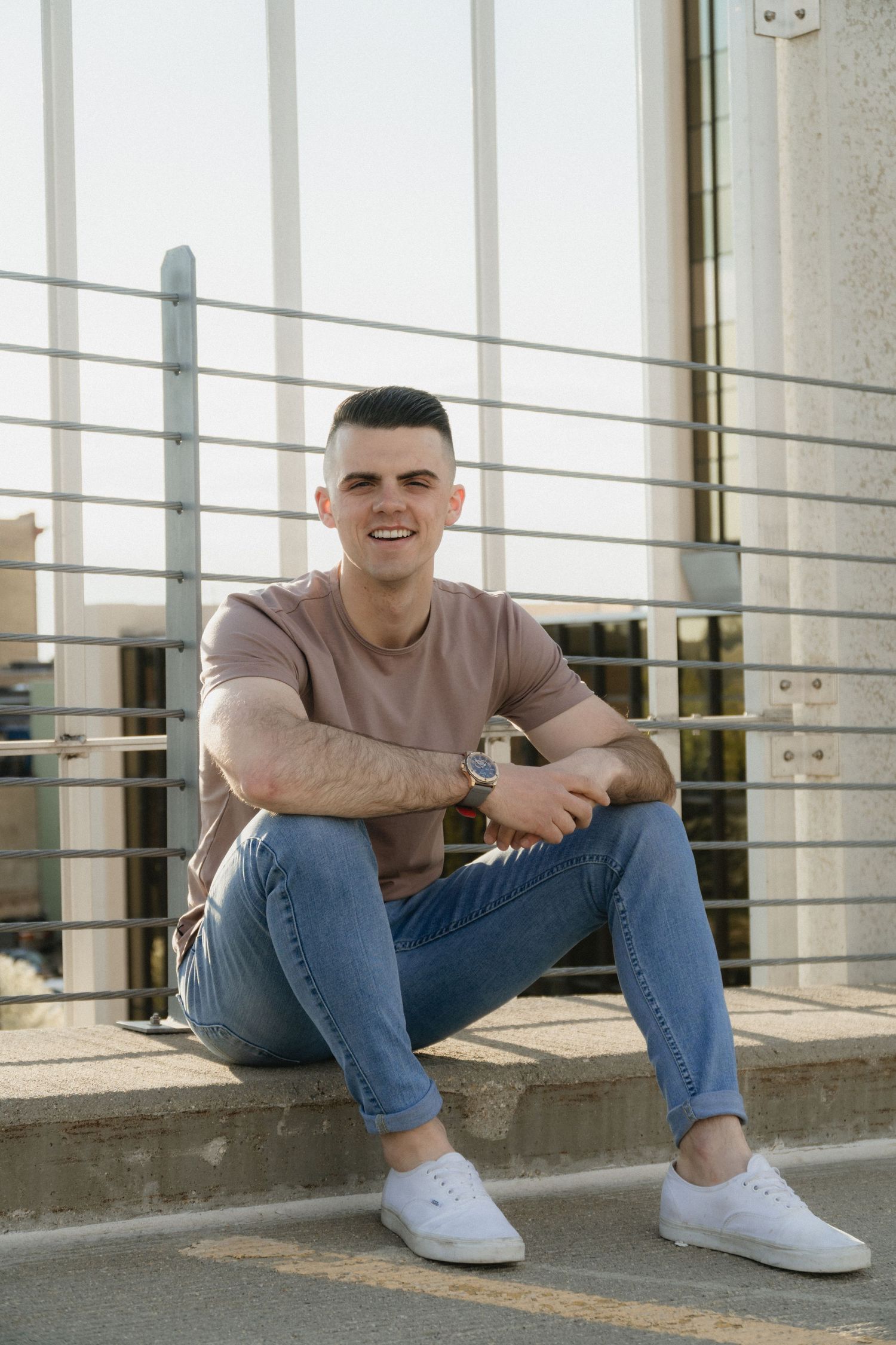 Young man in pink t-shirt and jeans sitting on steps outdoors, smiling confidently in natural sunlight.