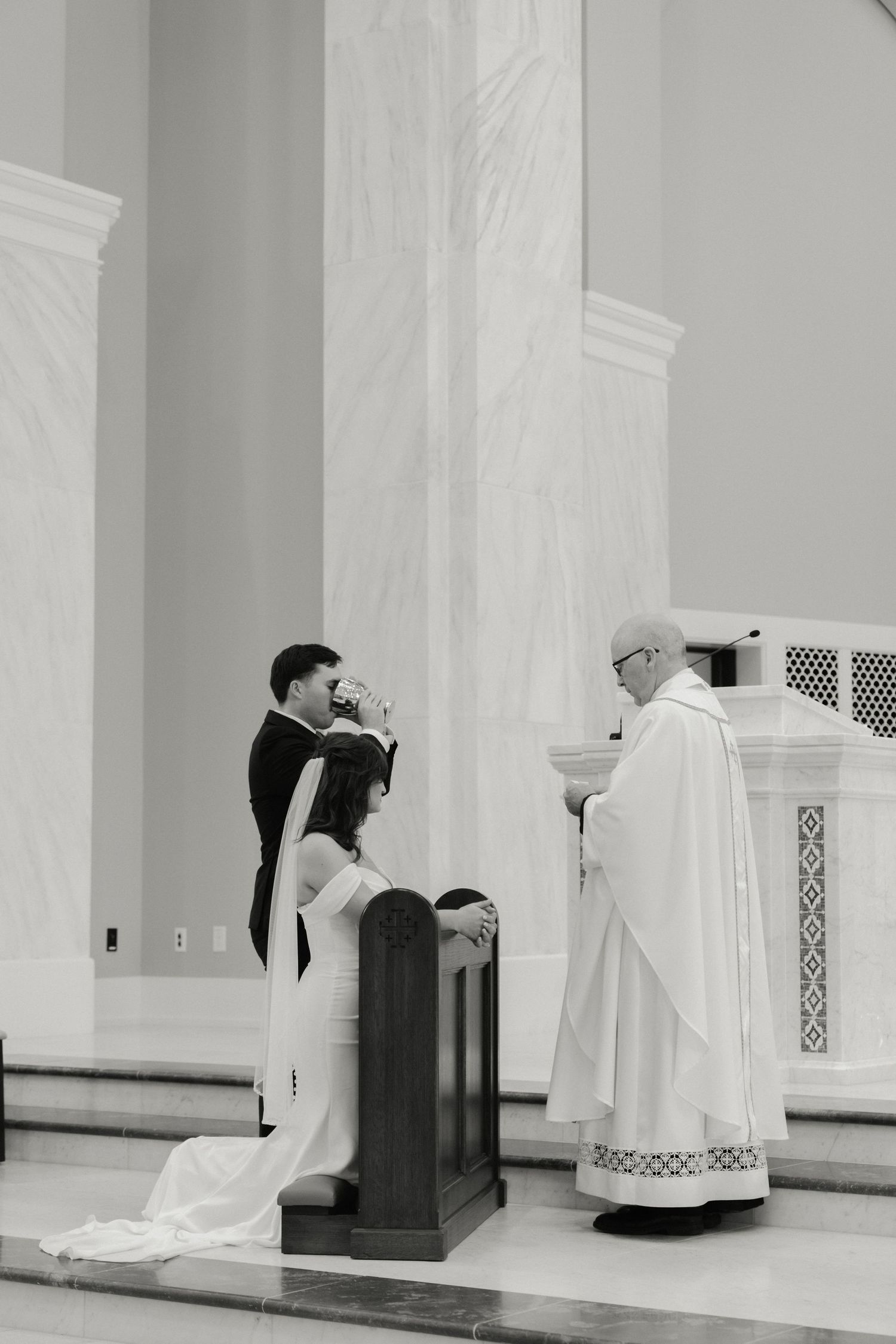 Black and white photo of a couple exchanging vows at a Catholic wedding ceremony with a priest officiating.