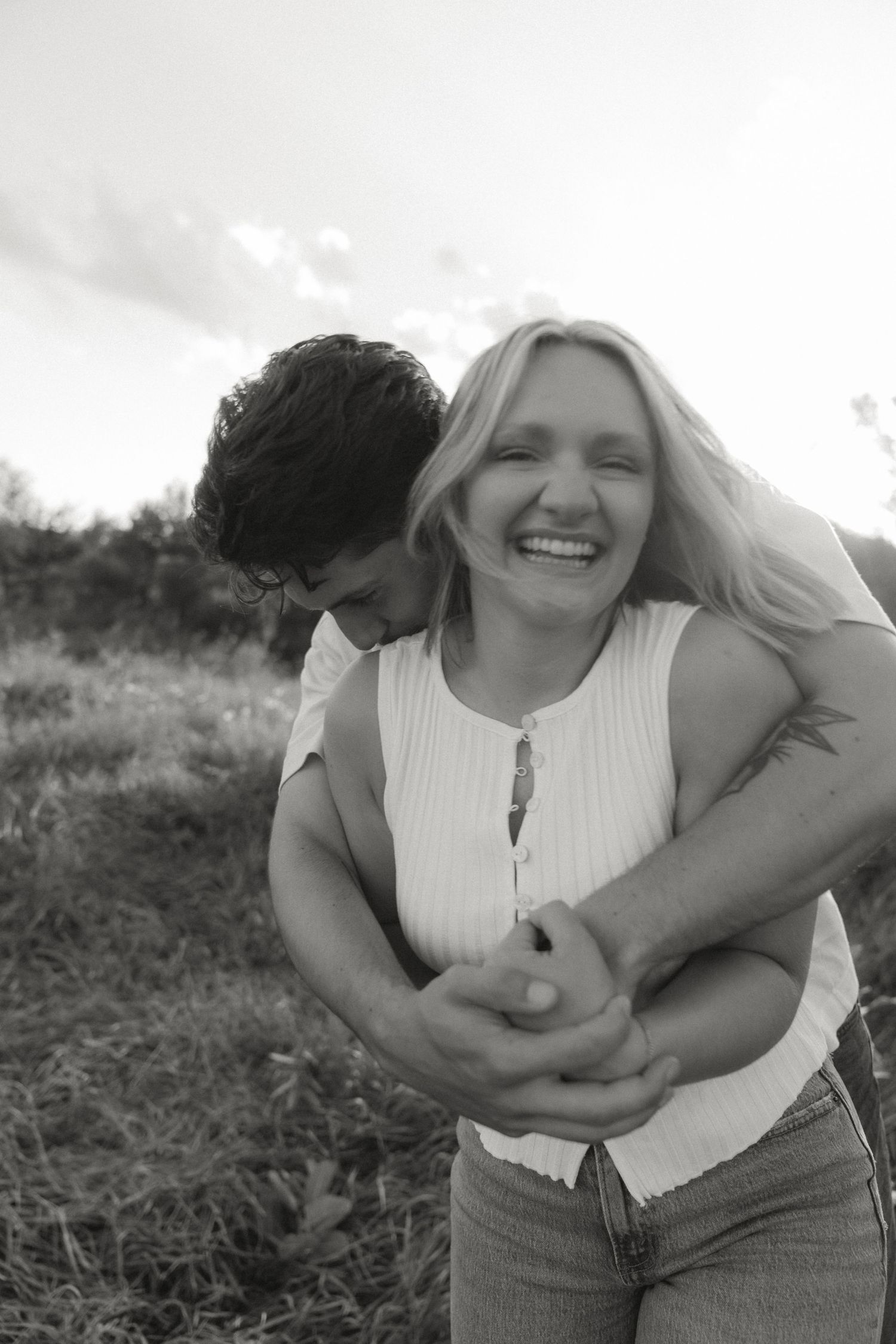 Black and white photo of a laughing blonde woman being hugged from behind by a dark-haired man outdoors.