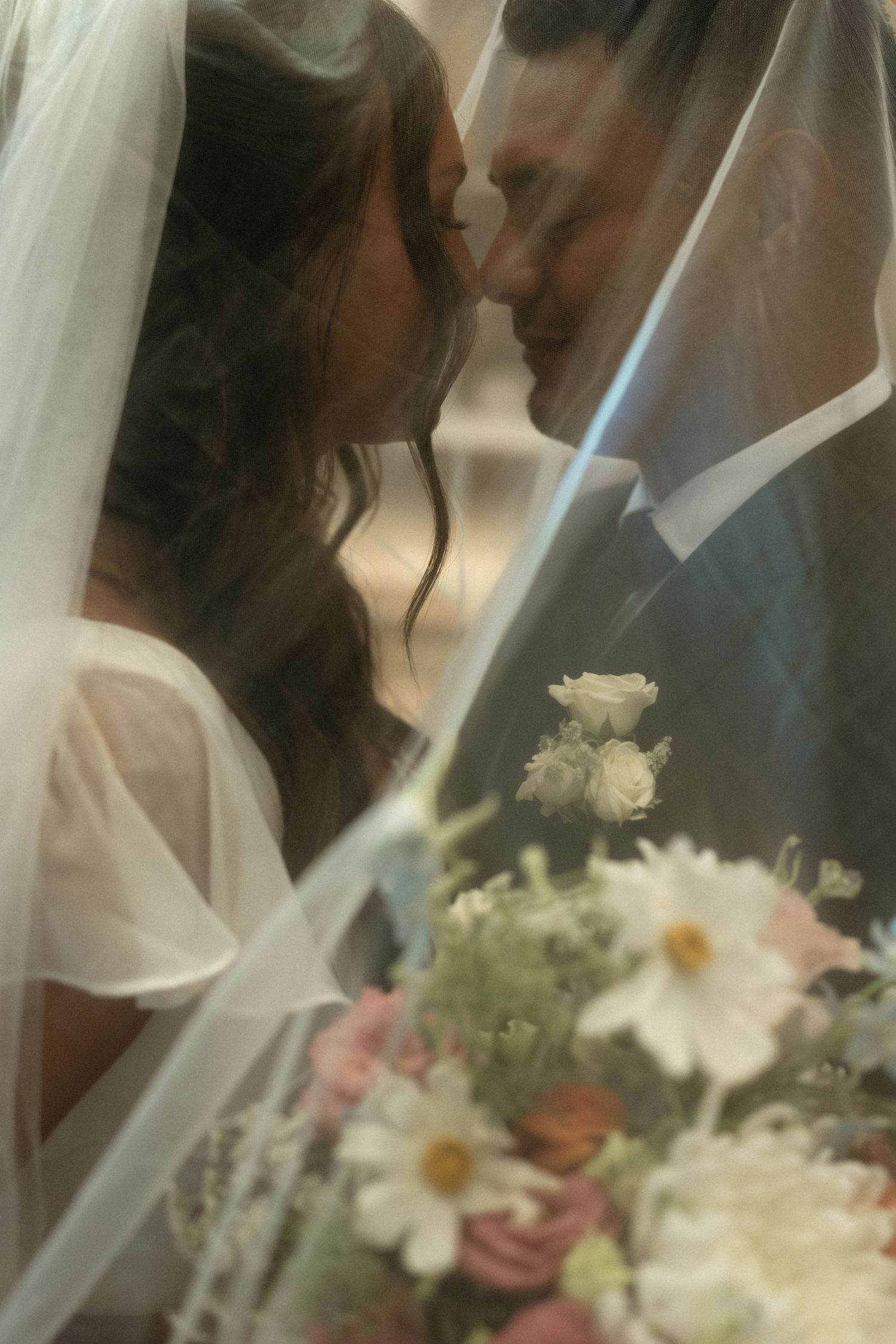 Bride and groom sharing a romantic kiss under a wedding veil with a beautiful floral bouquet in the foreground.