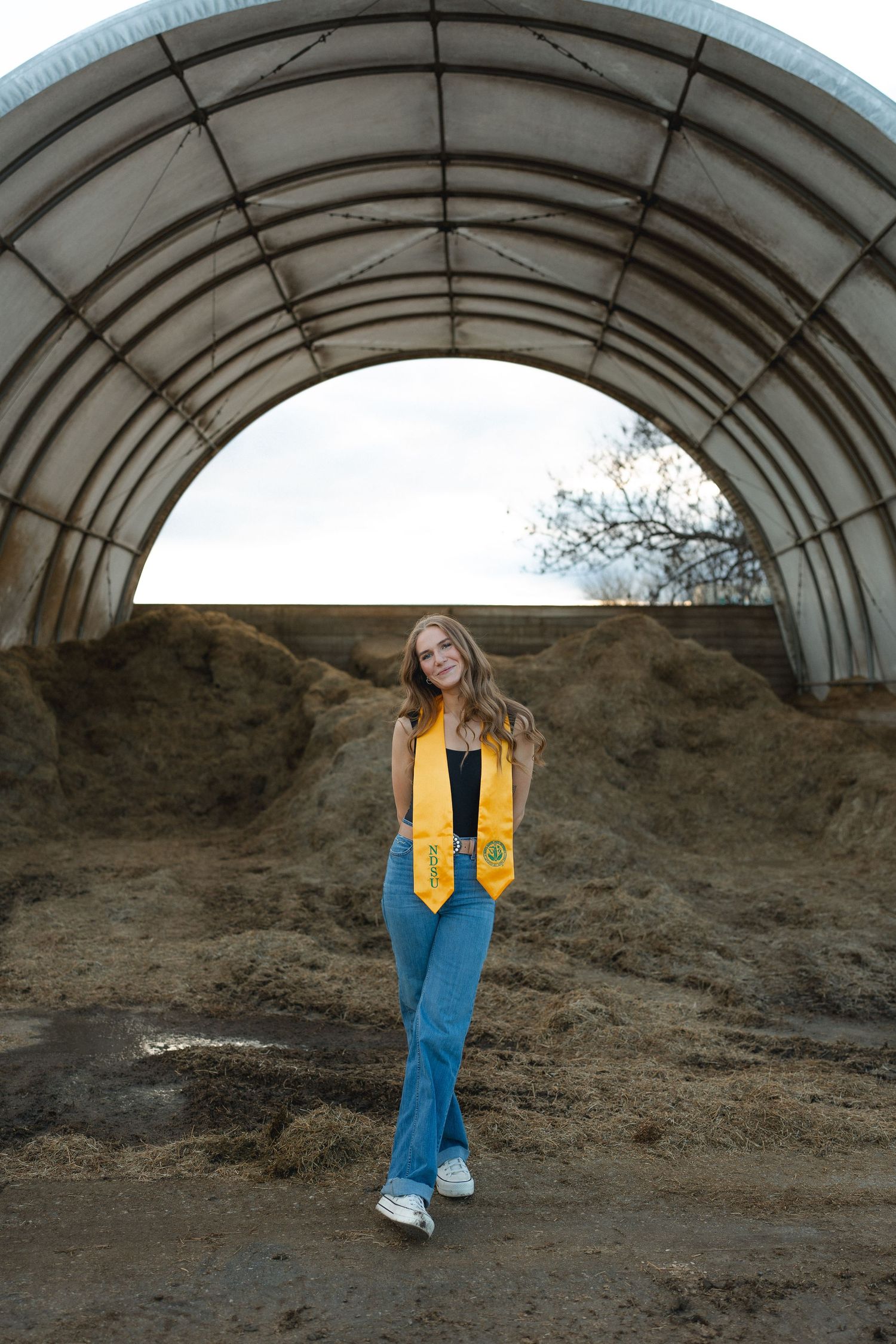 Young woman in graduation stole poses proudly under arched barn structure surrounded by hay bales.