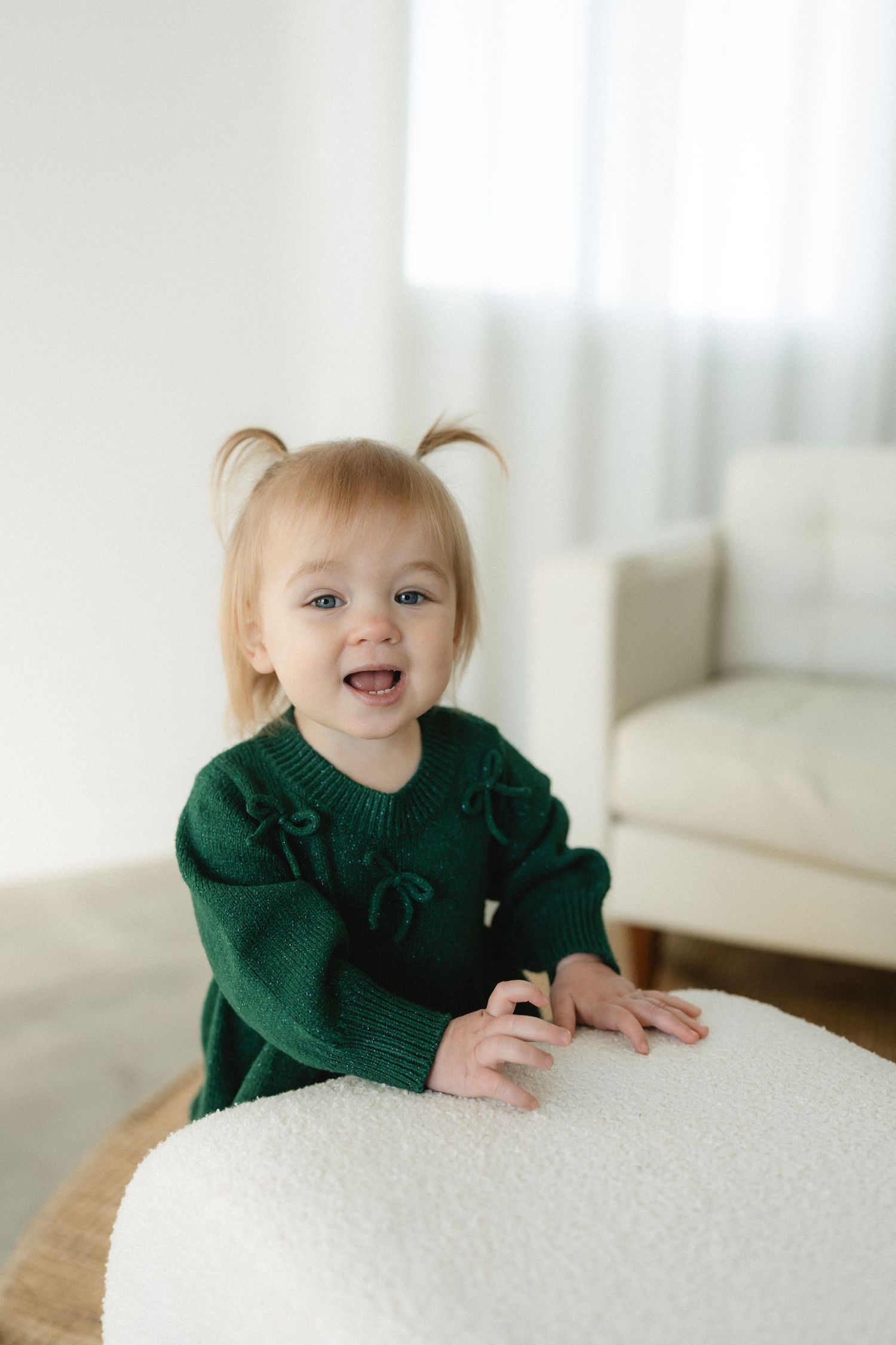 Cheerful toddler girl with pigtails wearing a green knit dress, smiling while leaning on a white ottoman.