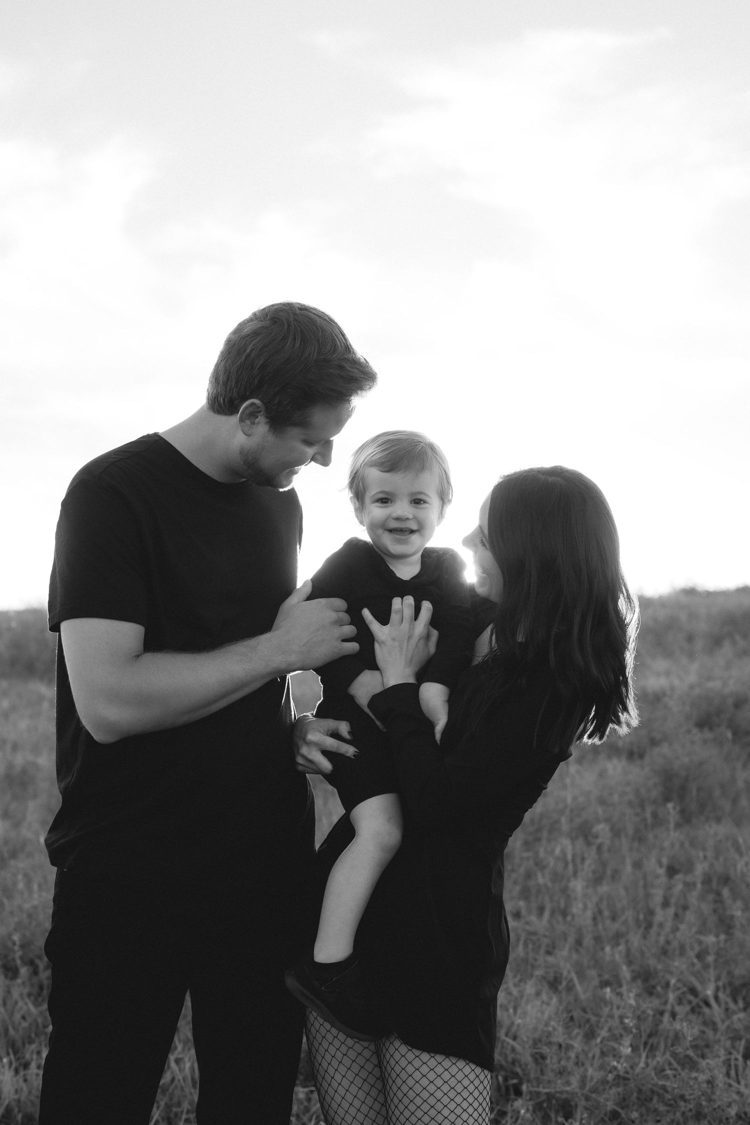 Black and white family photo of parents holding smiling toddler in field, all dressed in black.