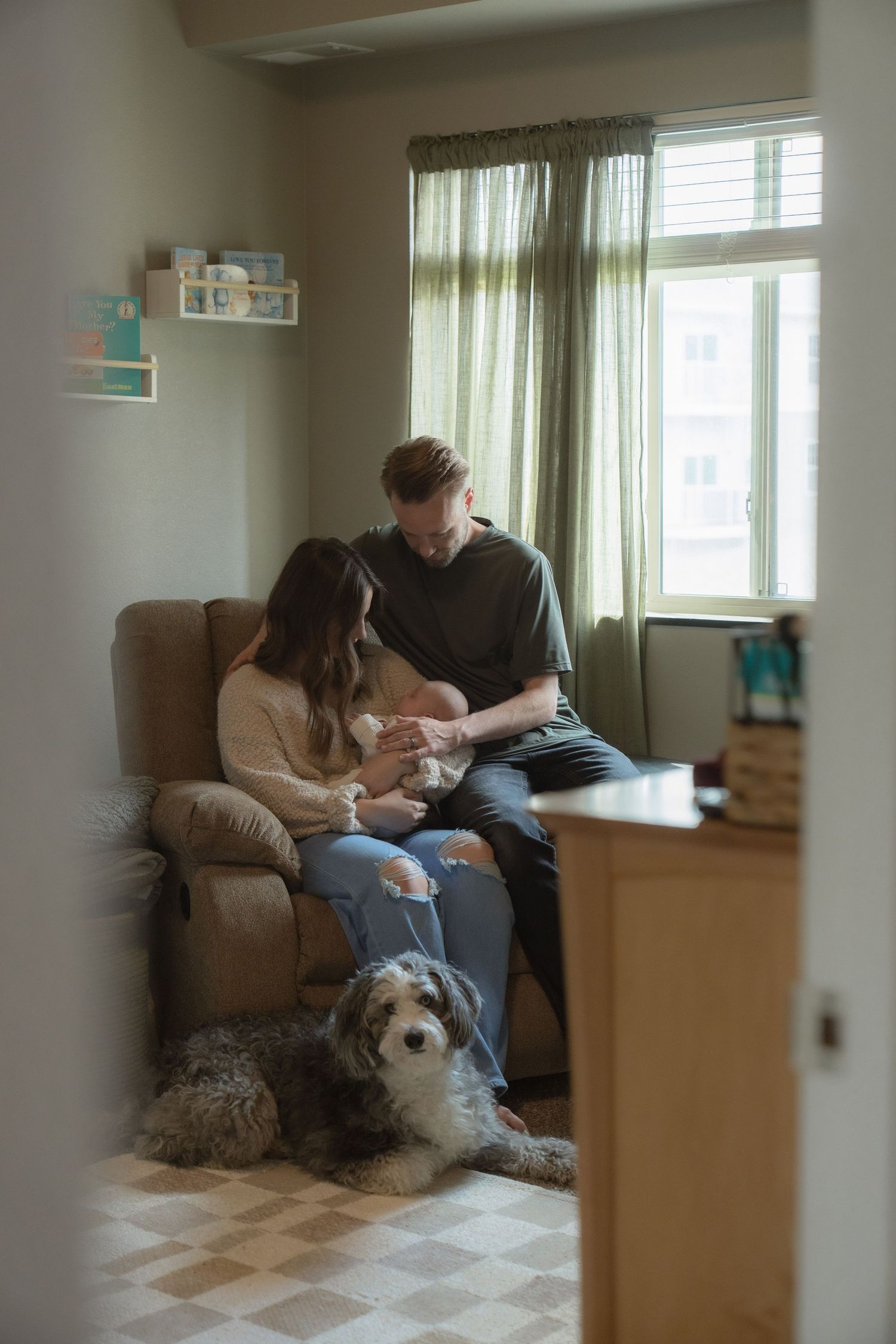 A couple sits in a recliner chair with their newborn baby while a fluffy dog rests on the floor beside them.
