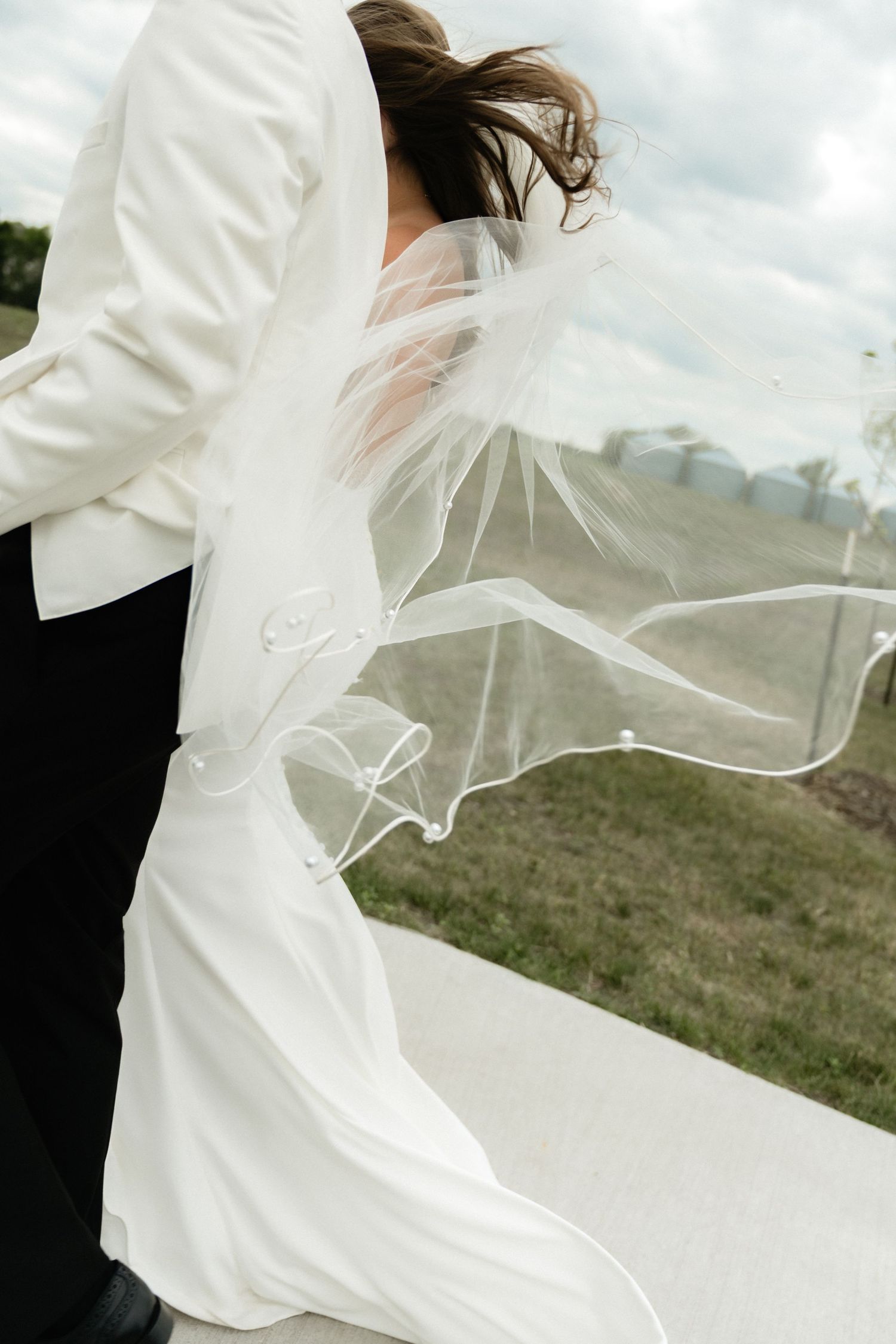 Bride in white gown and flowing veil blowing in wind, embracing groom in black suit outdoors on cloudy day.