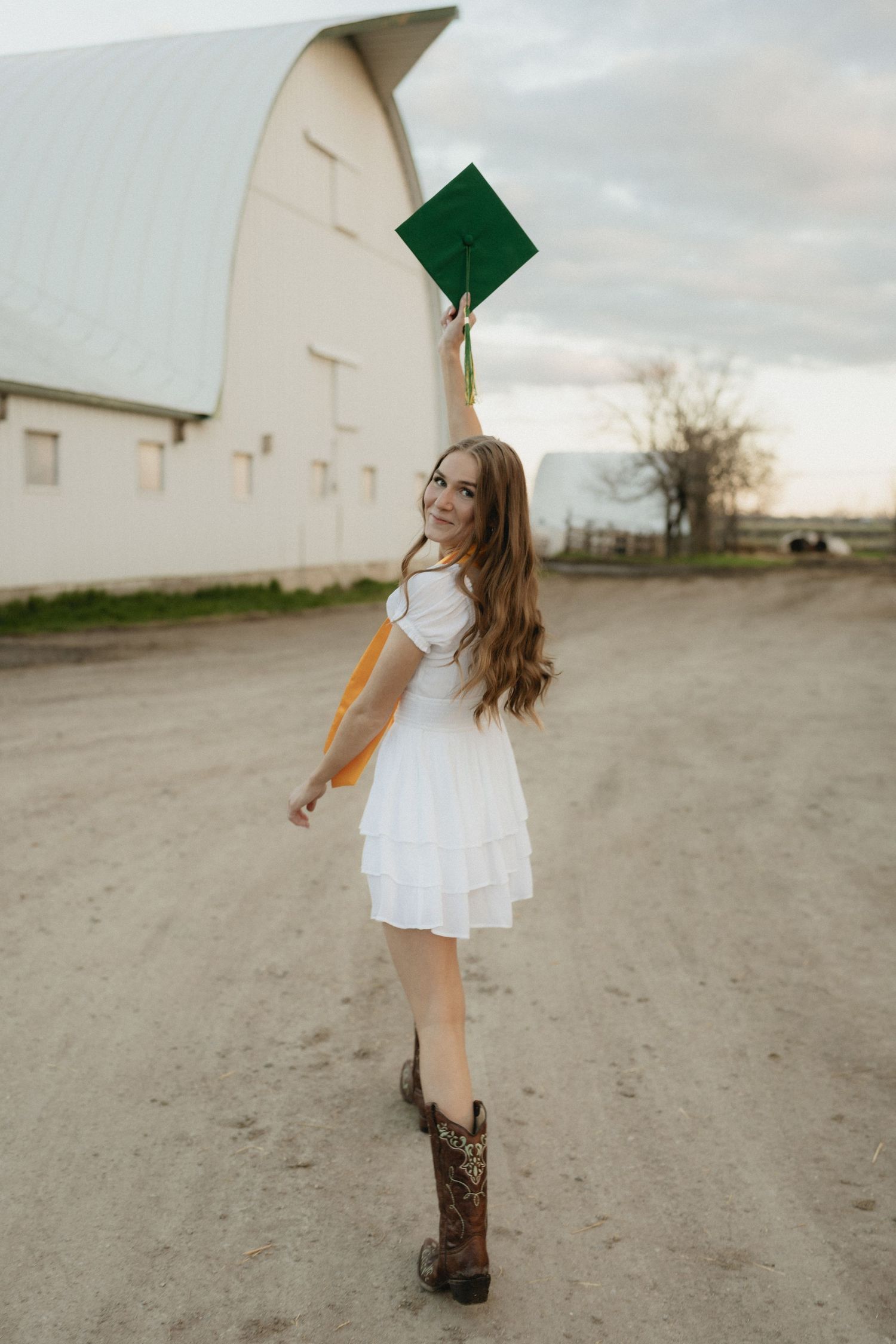 Young woman in white dress and cowboy boots holds green graduation cap aloft on a dirt road near a white barn.