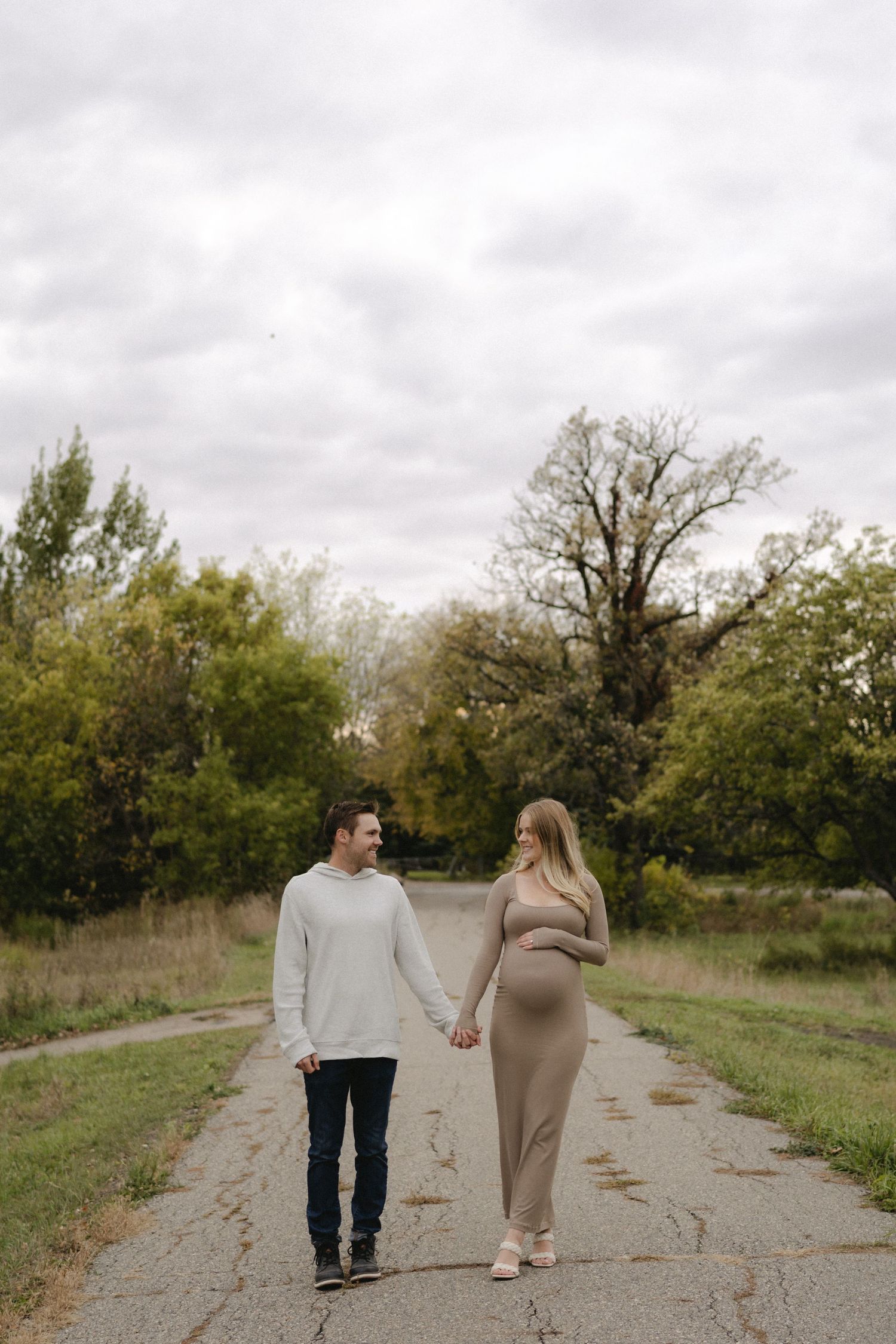 Pregnant couple holding hands walking on a path surrounded by autumn trees during an outdoor maternity photoshoot.