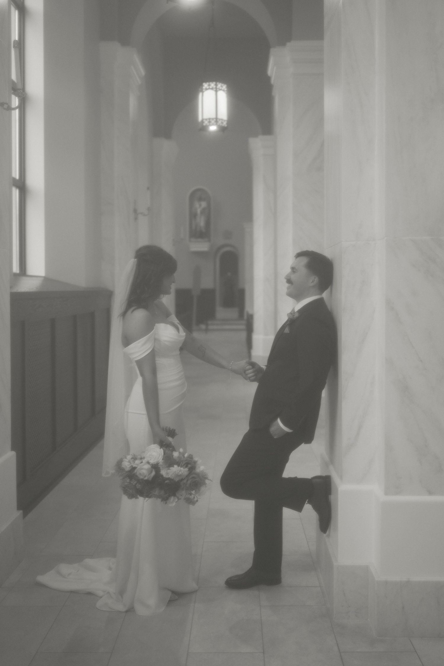 Black and white photo of bride holding bouquet and groom leaning against marble column in elegant church hallway.