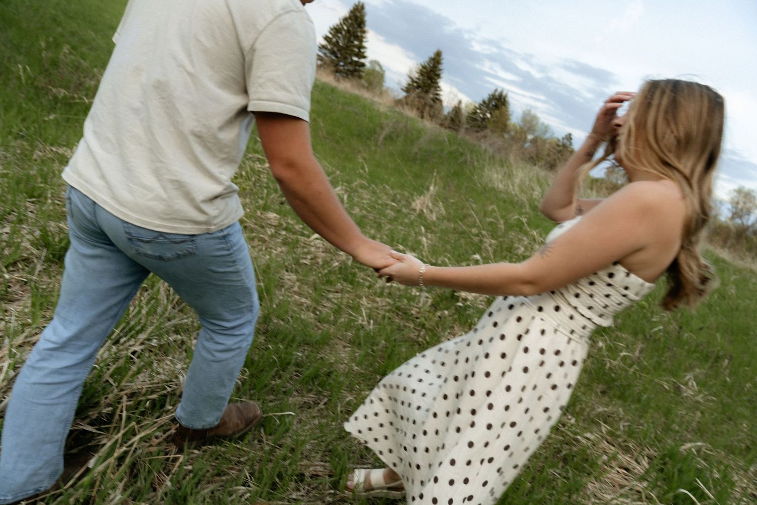Couple holding hands in a grassy field, woman in polka dot dress leaning back playfully on a cloudy day.