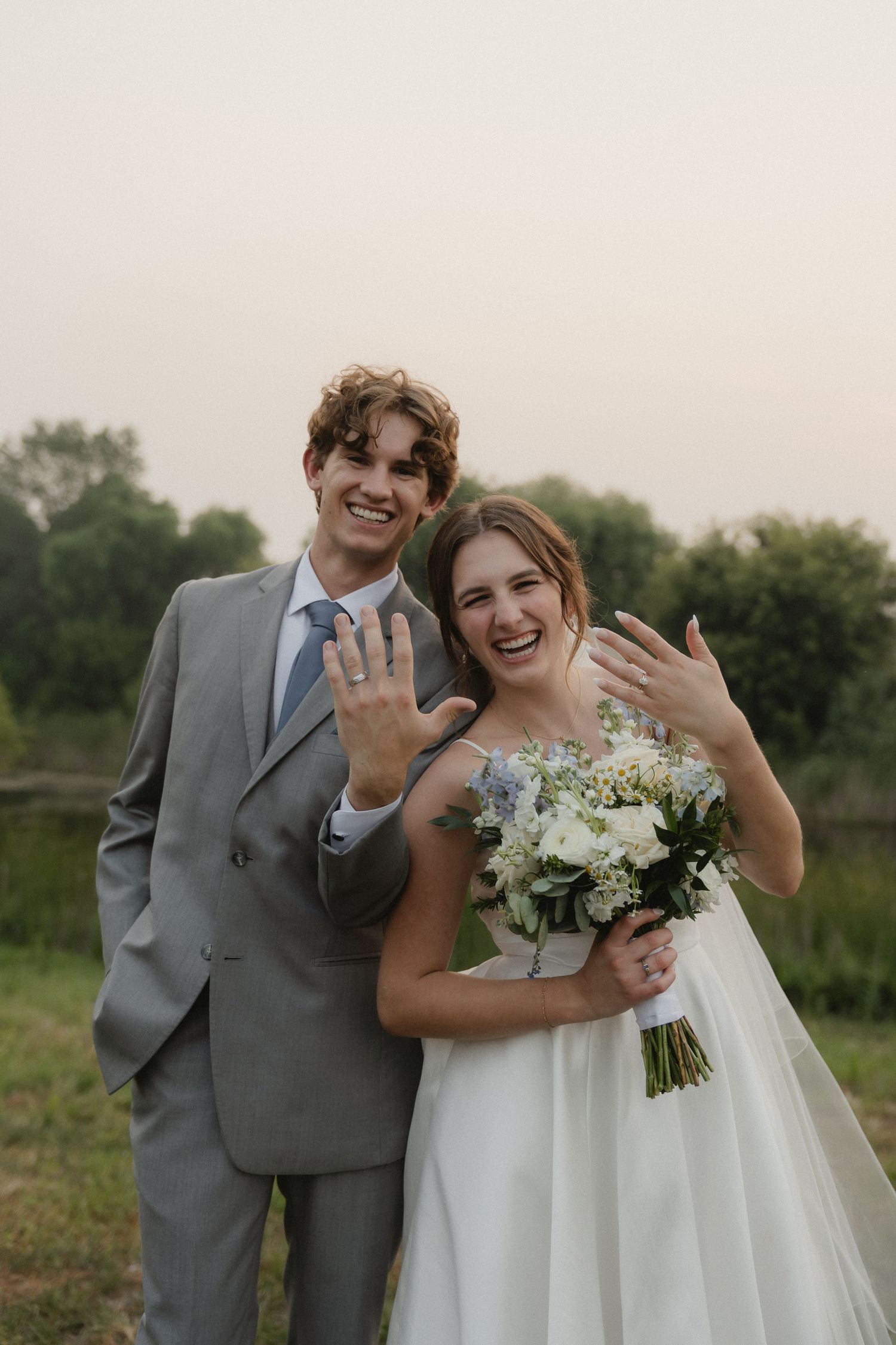 Happy newlyweds showing excitement while holding a white floral bouquet in an outdoor field.