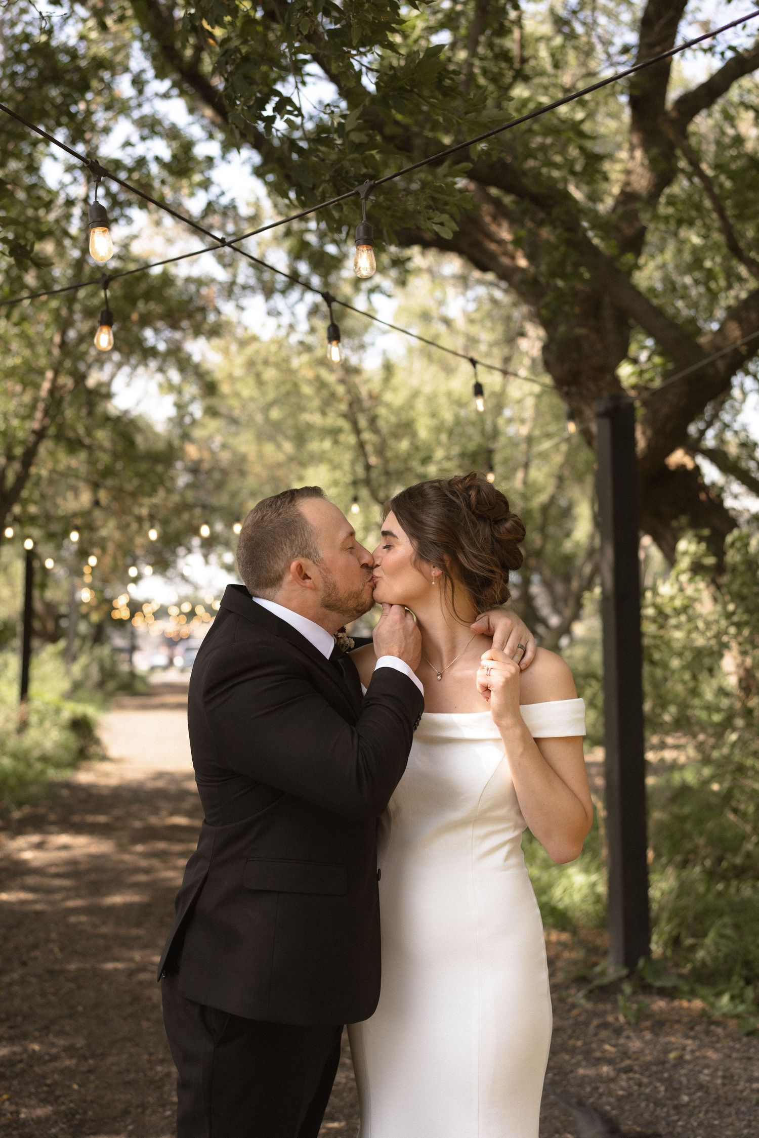 Bride and groom share a romantic kiss outdoors under string lights and lush green trees in wedding attire.