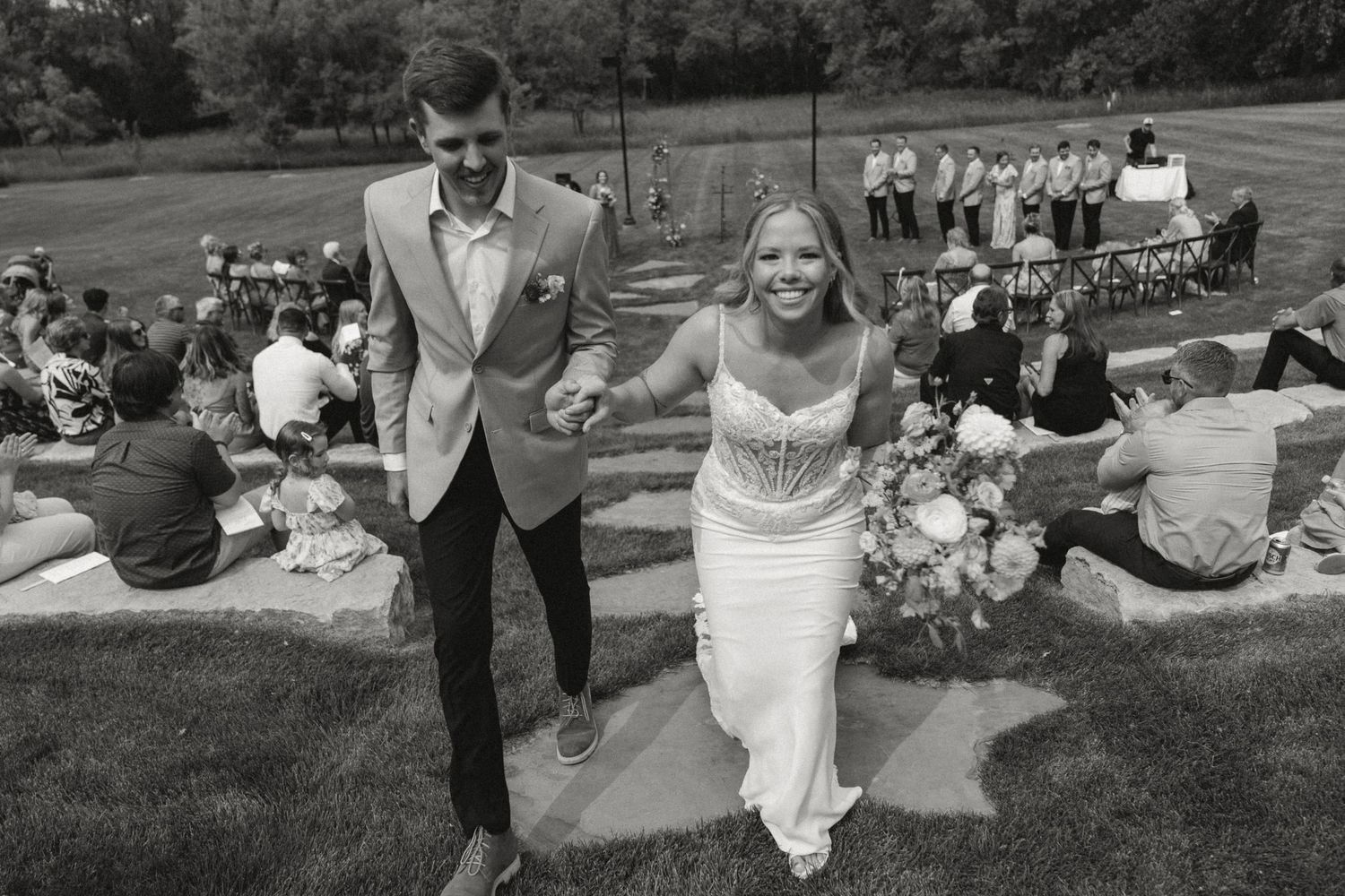 Black and white photo of a joyful newlywed couple walking hand in hand after their outdoor wedding ceremony.