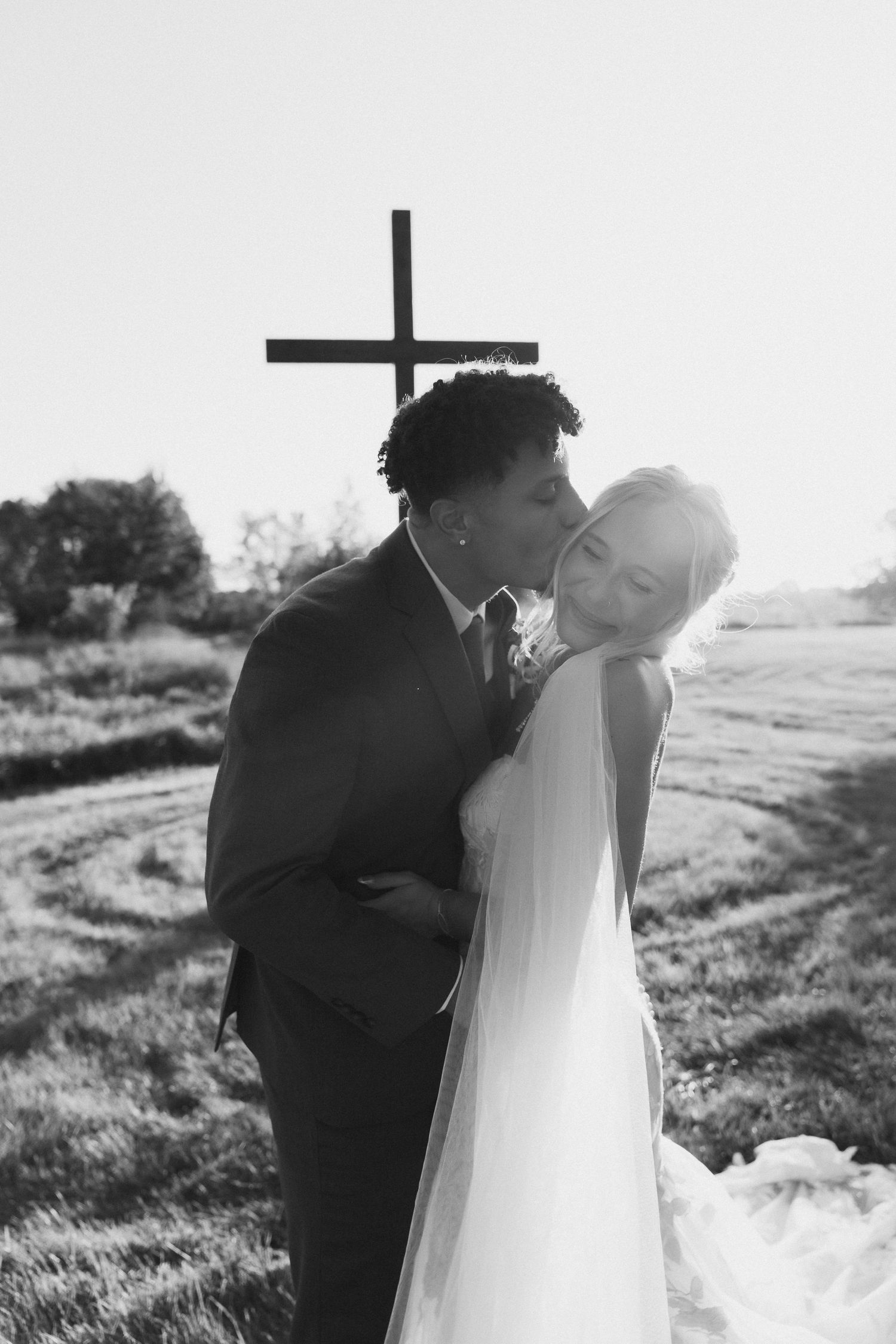 Black and white photo of a groom kissing bride on cheek outdoors with a wooden cross in background.