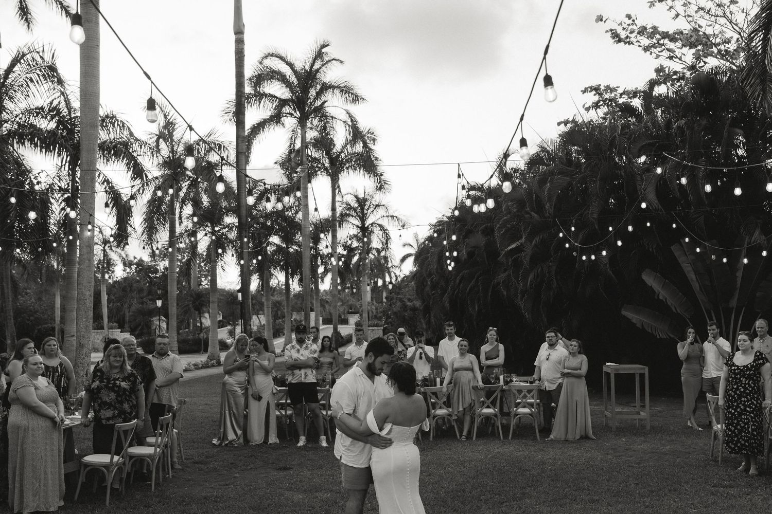 Couple shares first dance at outdoor wedding reception under string lights surrounded by palm trees and guests.