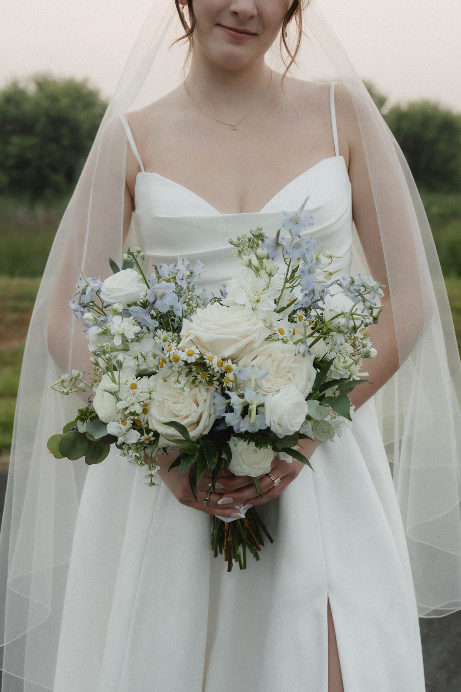 A delicate bridal bouquet features white roses and blue flowers with a flowing veil in the background.