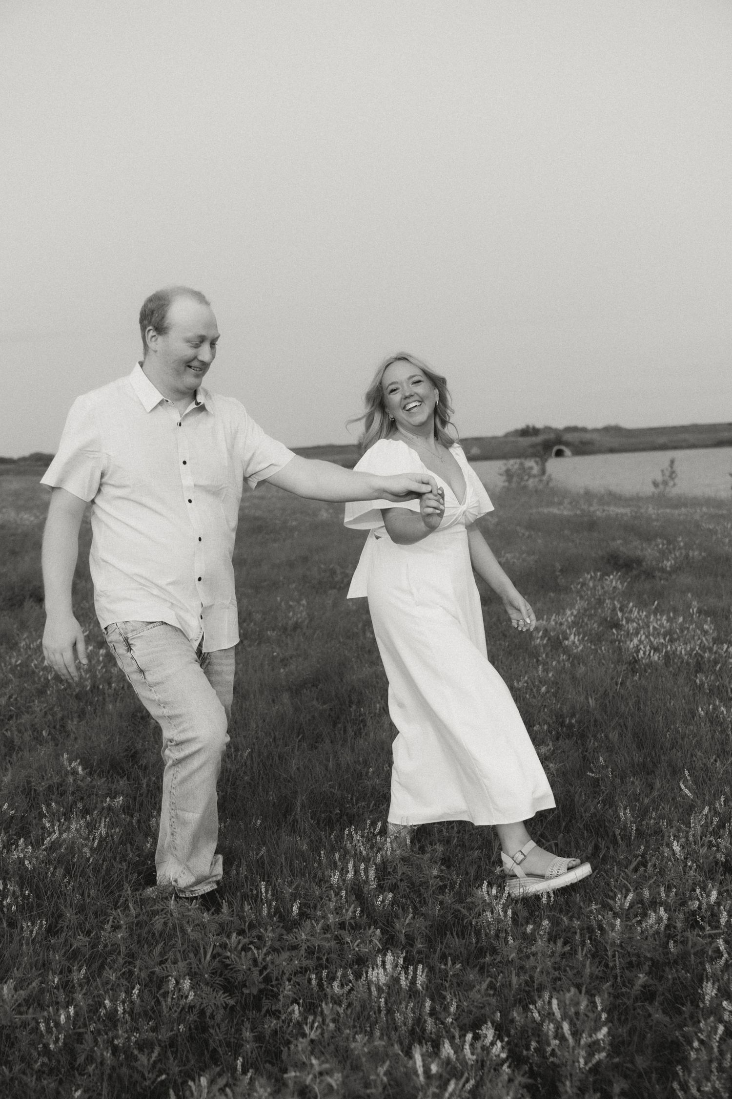 A couple in light colored clothing walks together in a grassy field.