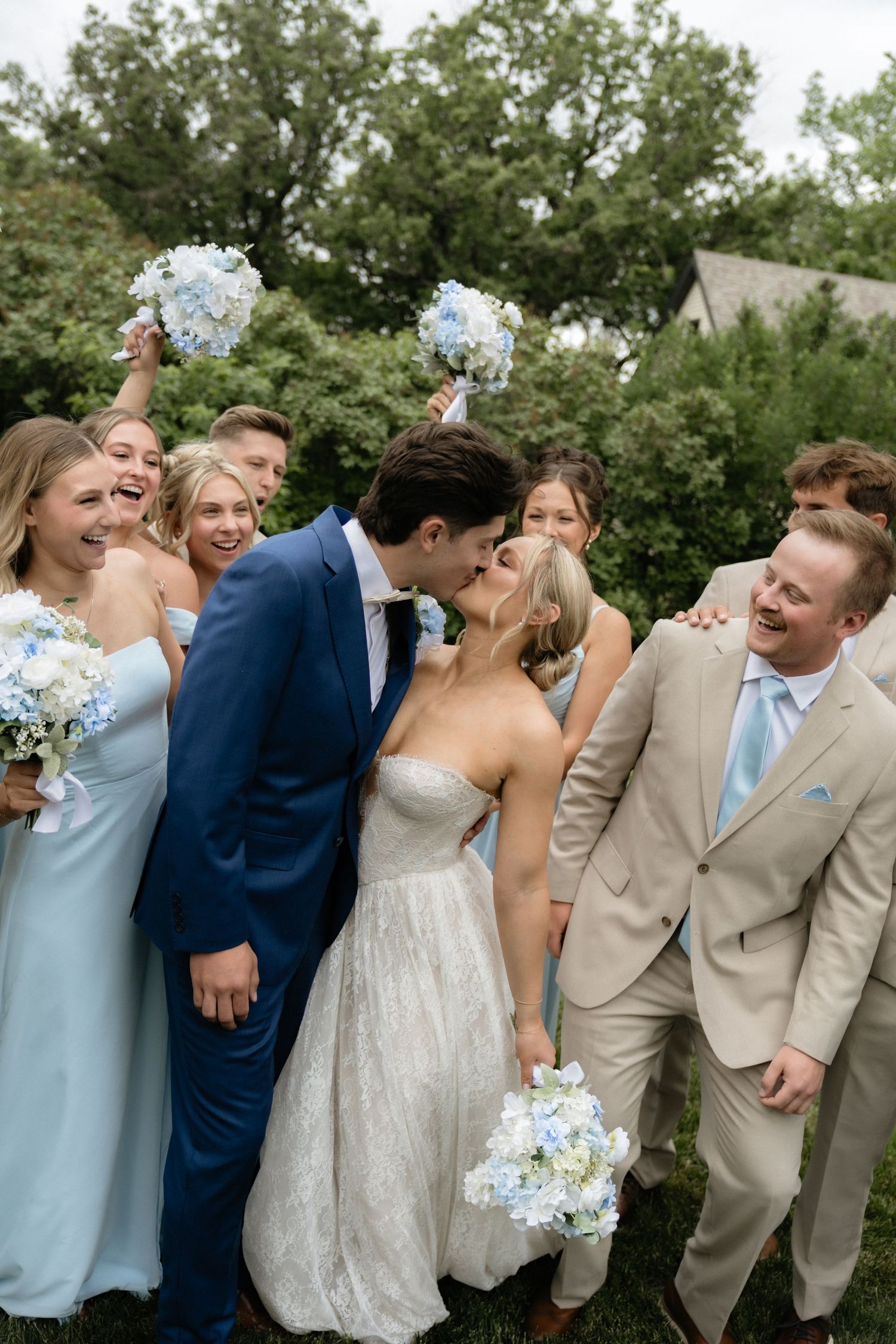 Bride and groom kiss surrounded by cheering wedding party in blue and tan attire holding light blue bouquets outdoors.