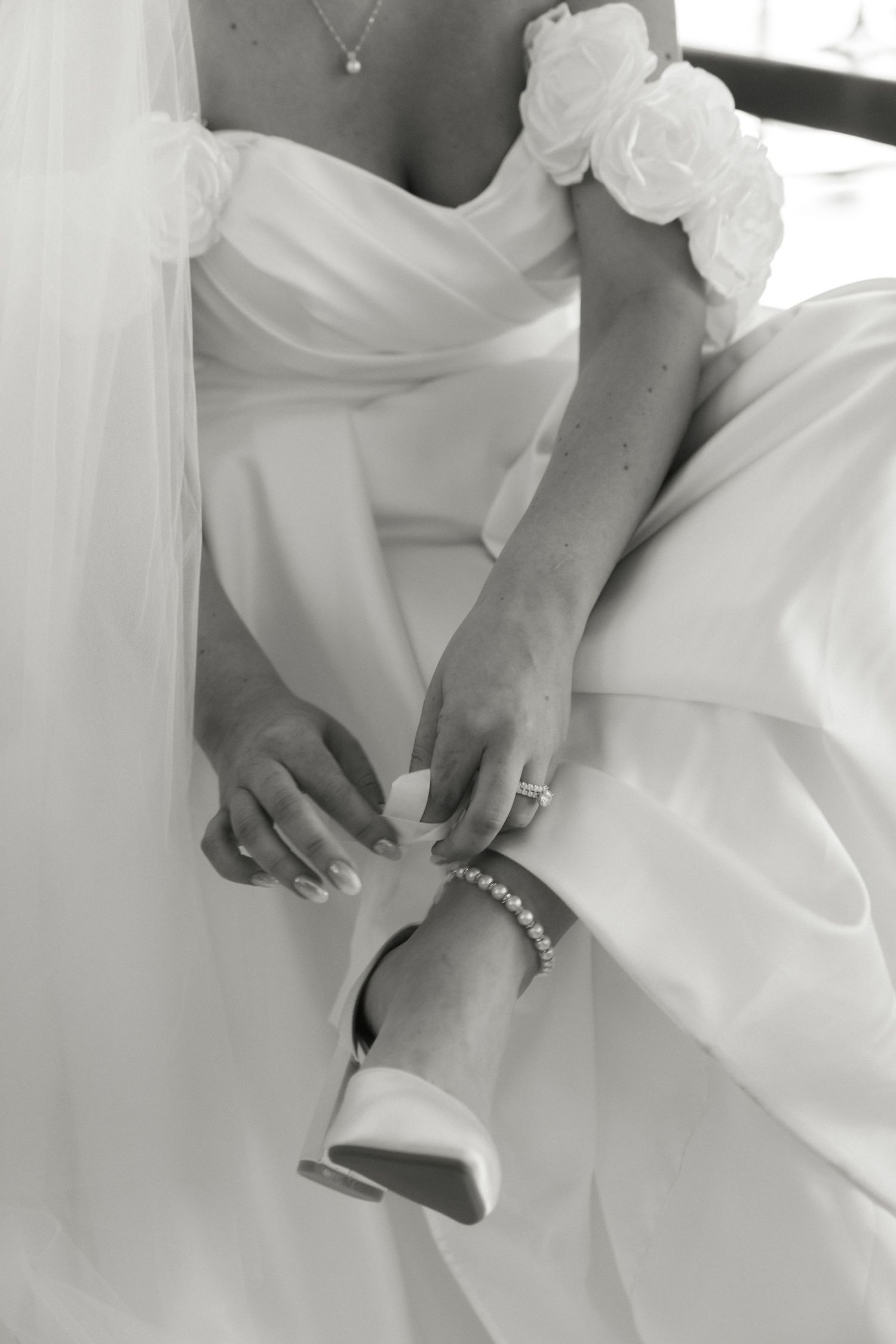 Bride in white wedding gown adjusting her shoe, wearing pearl bracelet and ring, black and white photo.
