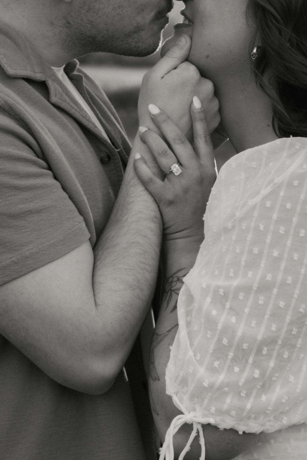 Black and white photo of a couple kissing while holding hands, showcasing an engagement ring on the woman's finger.