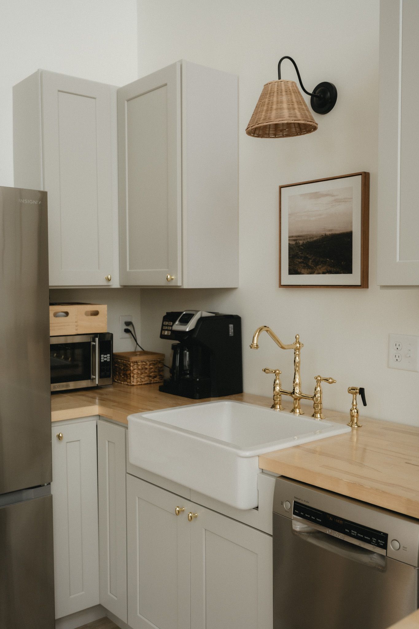White farmhouse kitchen with brass faucet, apron sink, butcher block counters, rattan sconce, and stainless appliances.